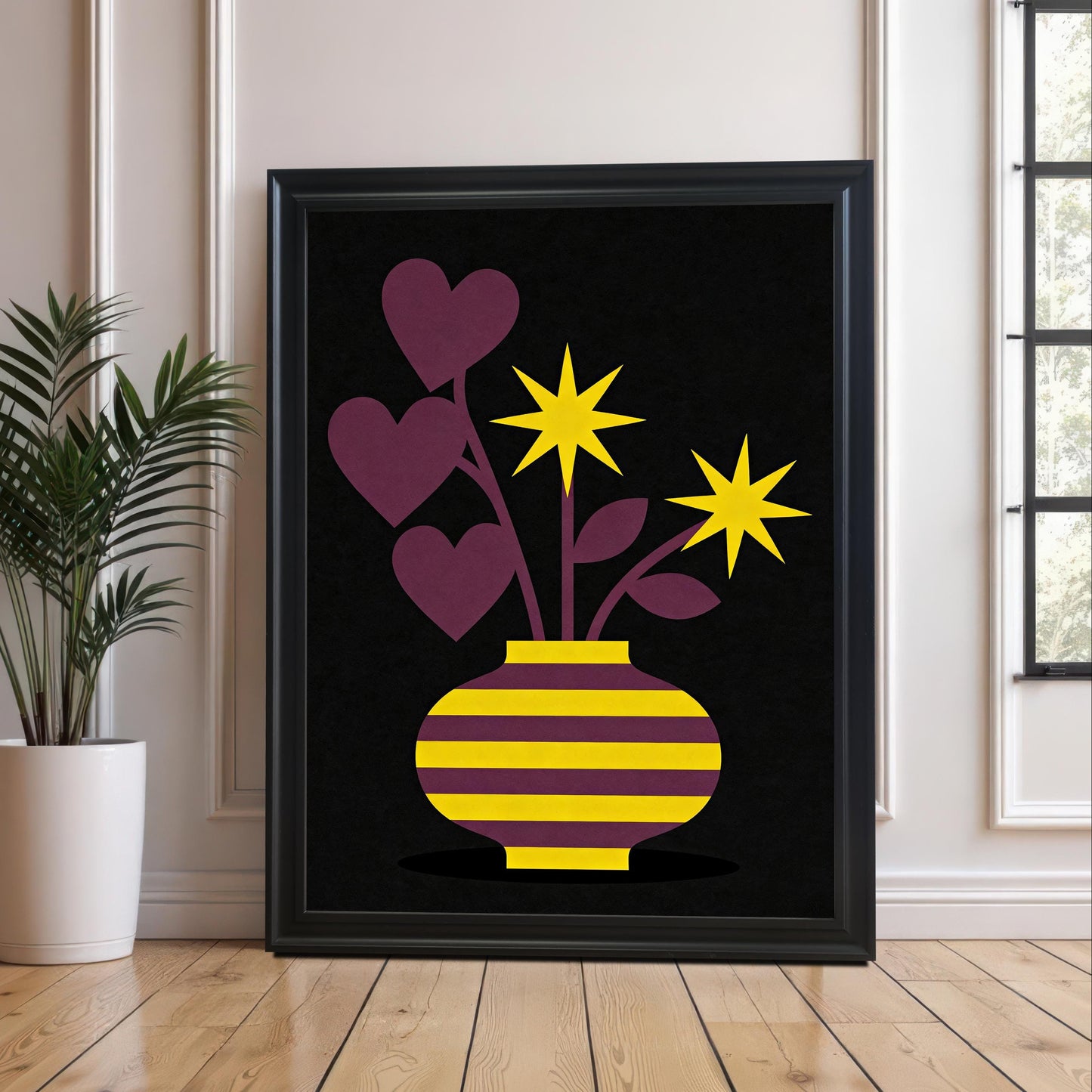 A large Black framed art print featuring a yellow and purple striped vase with purple heart-shaped leaves and bright yellow star flowers on a black background. The print rests on a wooden floor against a white paneled wall next to a tall houseplant in a white pot.