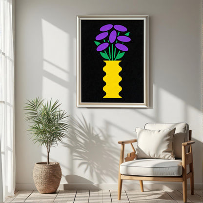 A large White framed art print featuring a bright yellow, wavy vase with stylized purple and green flowers on a black background, hanging on a white wall in a sunlit room. The print is positioned above a light wood chair with a white cushion and a potted plant in a woven basket.