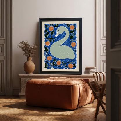 A framed print of the light gray swan and orange fruit artwork is displayed in a modern, sunlit room. The print is in a black frame, leaning against a mantlepiece above a large, square, burnt-orange leather ottoman.