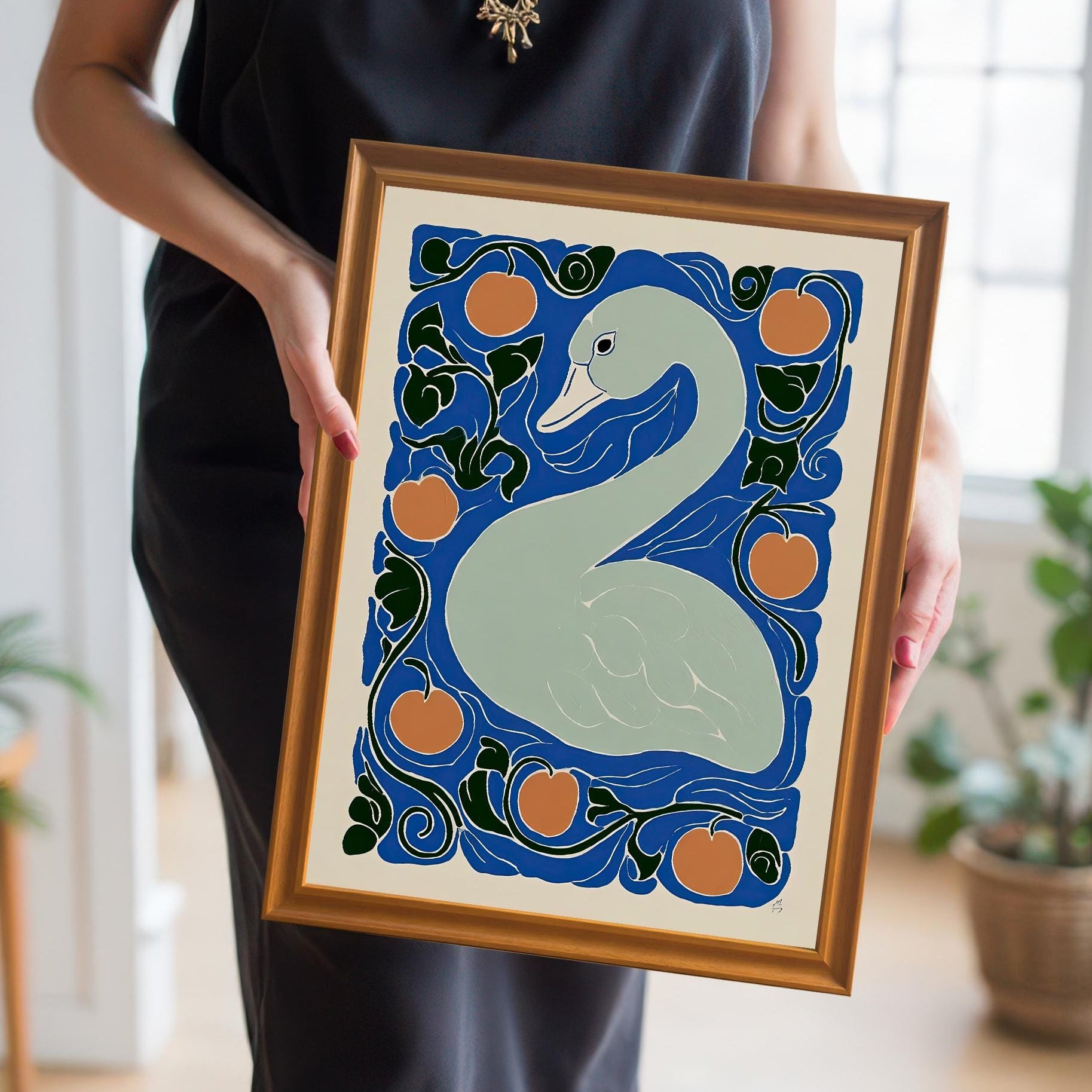 A person in a black dress holds a framed print featuring a light gray swan surrounded by orange circles (fruit) and dark blue and green swirling foliage, set against a royal blue background. The frame is a thin, shiny gold/bronze color.