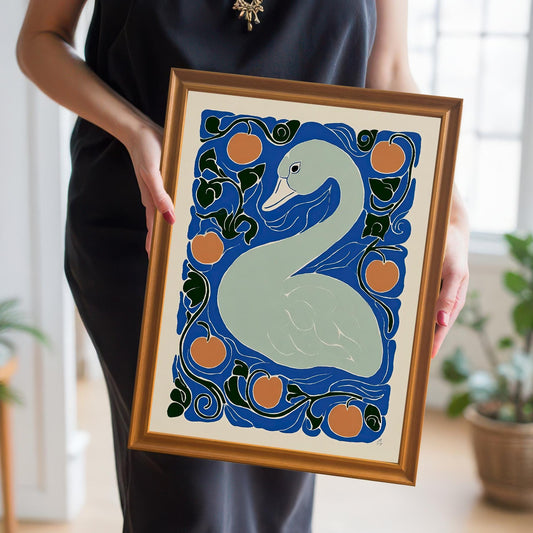 A person in a black dress holds a framed print featuring a light gray swan surrounded by orange circles (fruit) and dark blue and green swirling foliage, set against a royal blue background. The frame is a thin, shiny gold/bronze color.