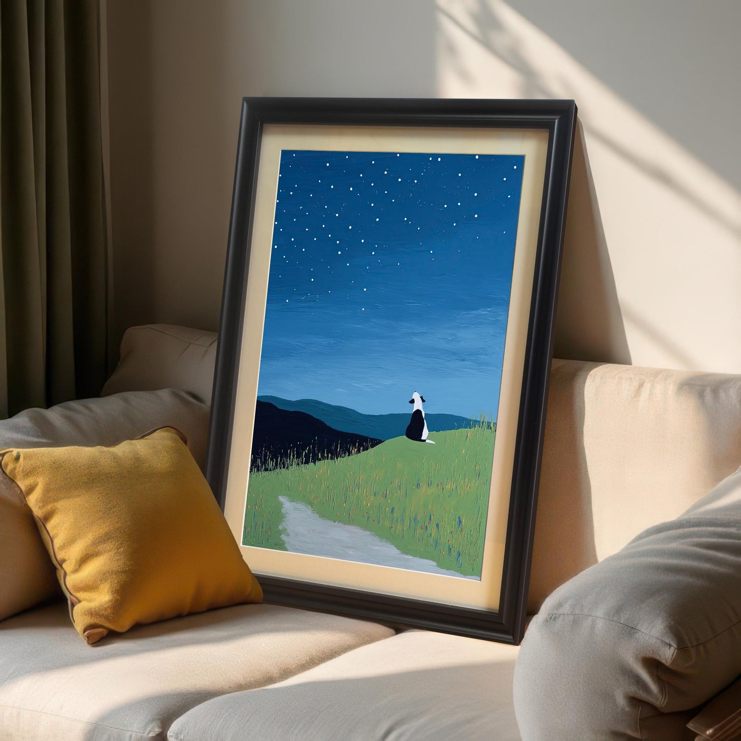 Close-up, angled view of the framed starry night dog print. The print is in a Black frame with a mount and is leaning on a beige sofa next to a yellow pillow, with dramatic sunlight shining in from the side.