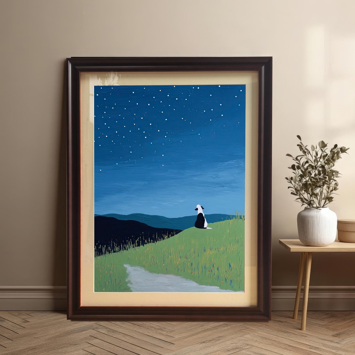Framed starry night dog print in a Dark Brown/Coffee frame with a mount, leaning on a wooden floor against a tan wall next to a small wooden side table with a white vase and dried plant.