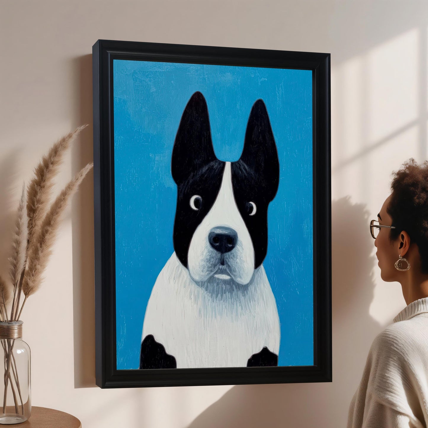 A painting of a black and white dog (resembling a Boston Terrier or French Bulldog) with large ears and wide eyes on a bright blue background, displayed in a Black frame hanging on a tan wall. A person wearing glasses looks closely at the framed art on the right.