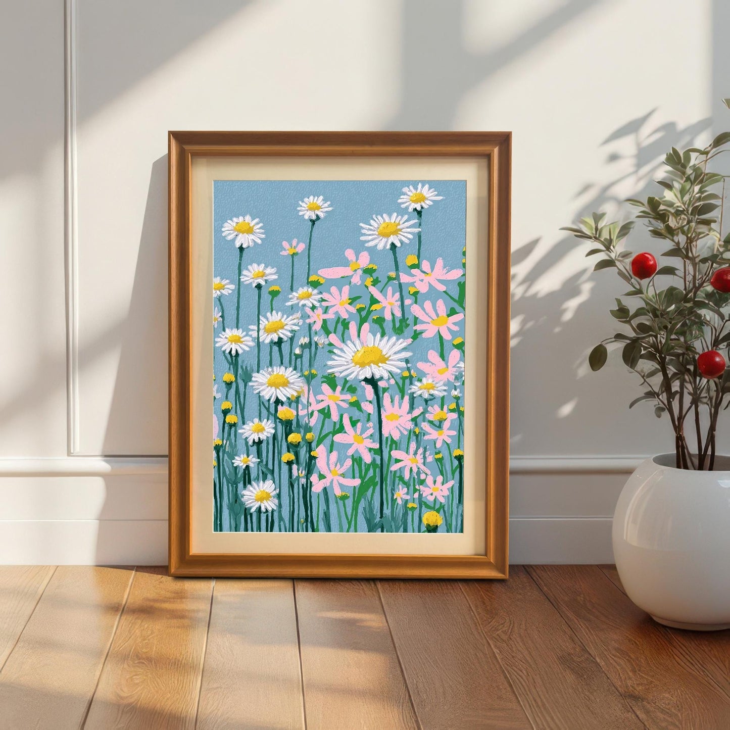 A framed art print featuring an illustration of white and pink daisies with yellow centers and stems against a light blue background. The print is in an Orange/Wood frame with a light-colored mat, leaning against a white wall next to a large white planter.