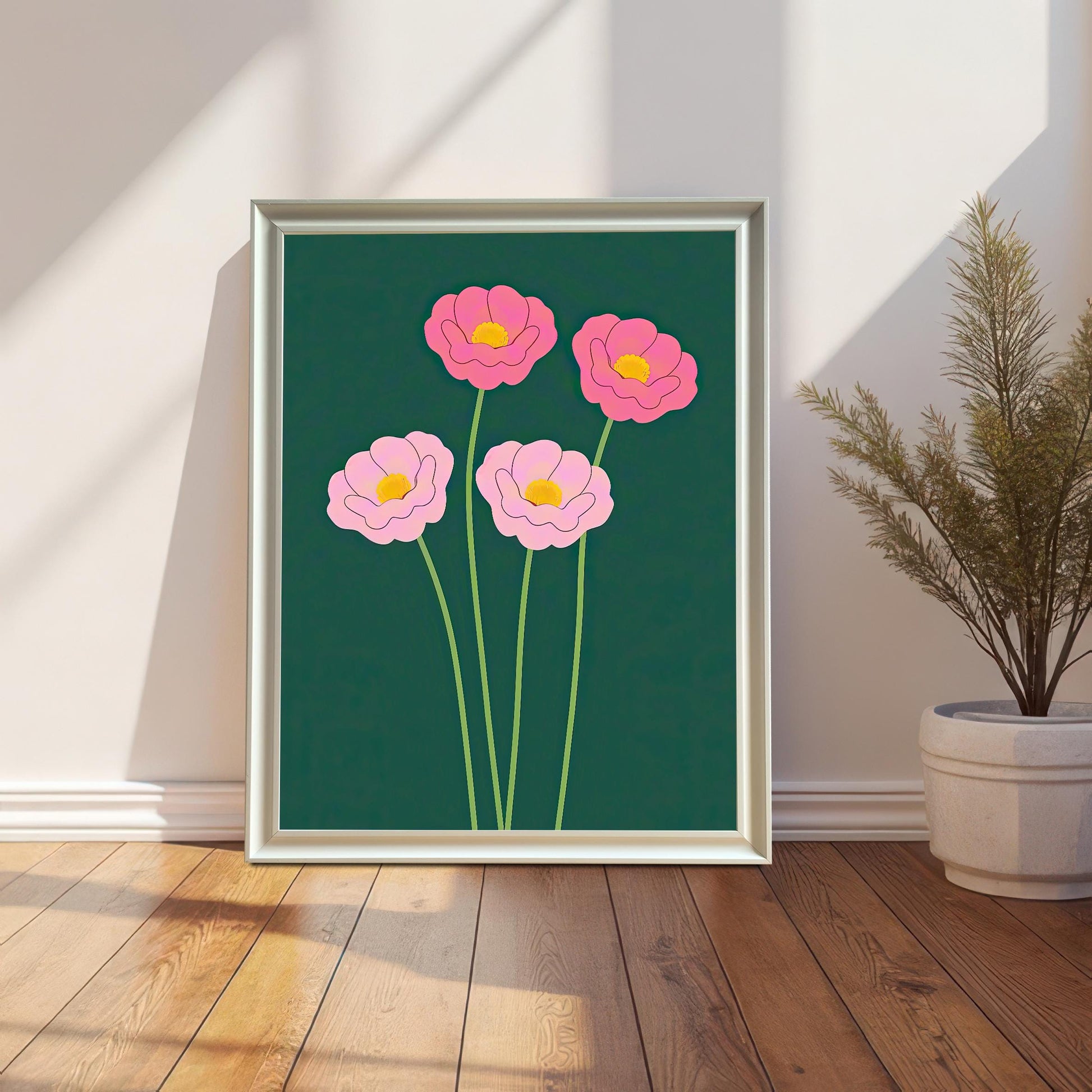 The art print featuring four stylized pink flowers on a dark forest green background, displayed in a White Frame. The print is leaning on a hardwood floor against a white wall with wainscoting, with a tall potted plant beside it.