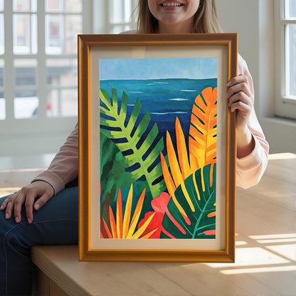 Lifestyle image of a smiling person sitting down and holding up the framed art print of the vibrant tropical leaves and blue ocean. The print is in a gold/Orange wood frame with a mount.