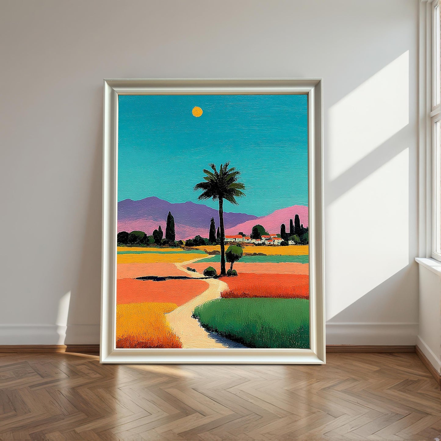 A large framed print of the vibrant, colorful landscape with a palm tree, in a white/silver frame, leaning on a wood floor by a sunlit window.
