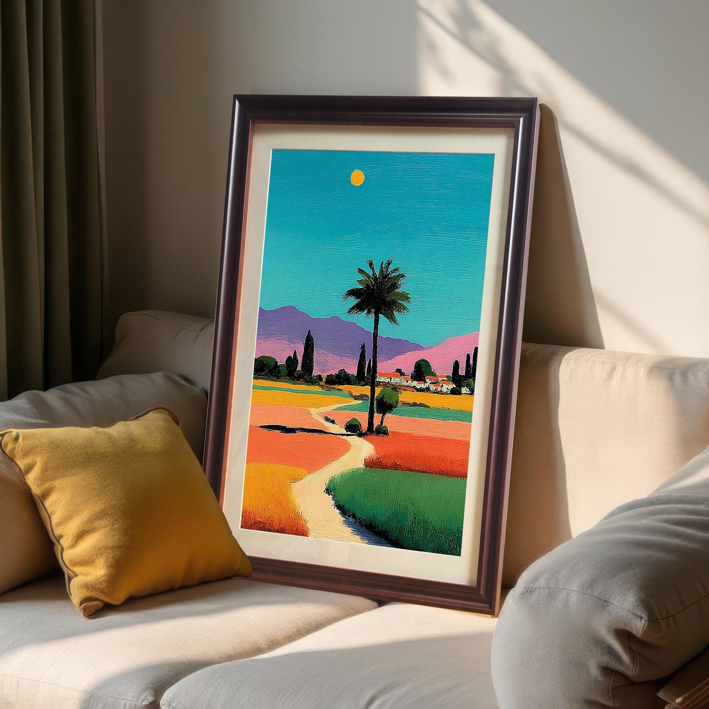 A framed print of the colorful palm tree landscape in a dark brown/coffee frame, leaning against a white couch next to a yellow pillow.