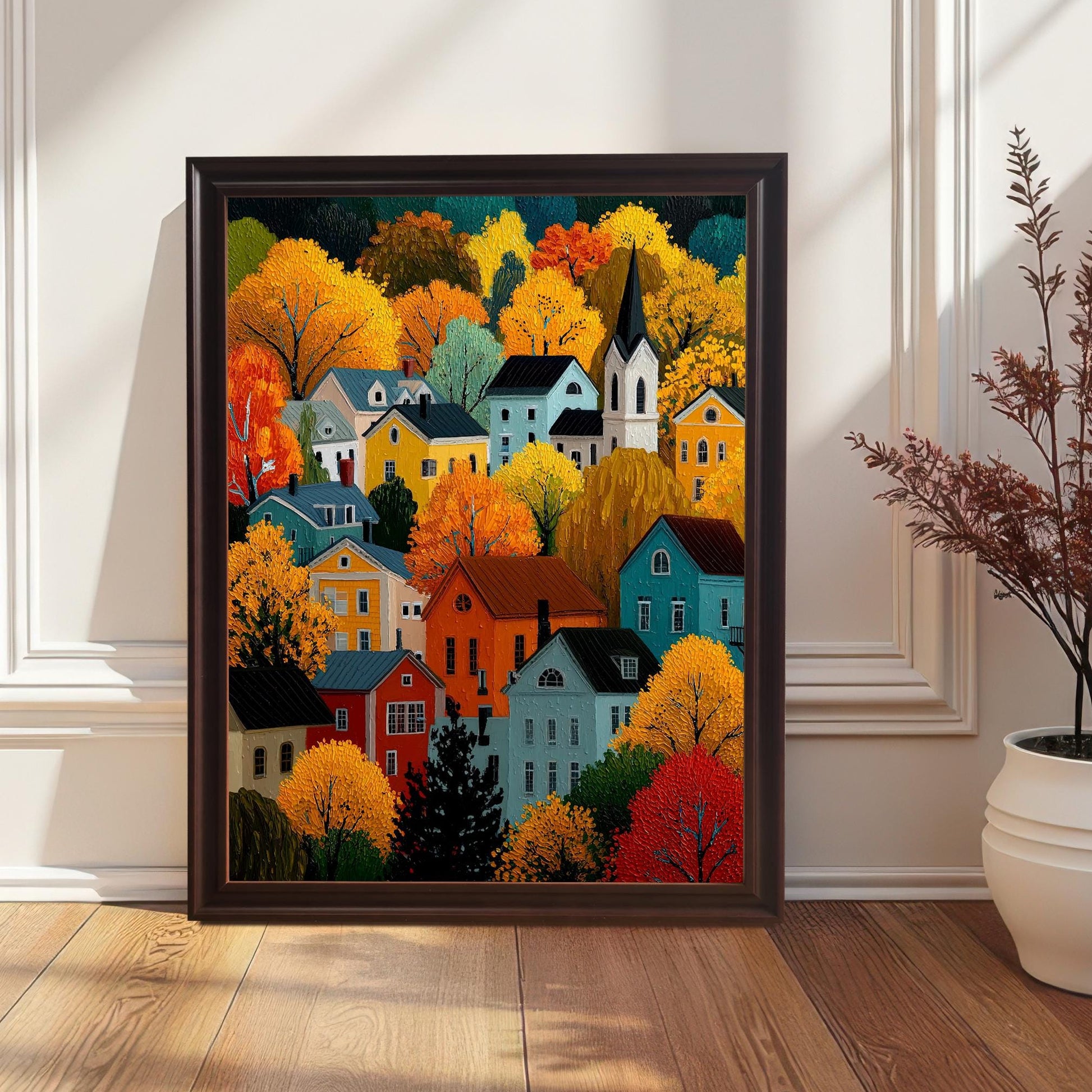 A framed print of a colorful autumn village scene, in a dark brown/coffee frame, leaning on a wooden floor against a white paneled wall near a potted plant.