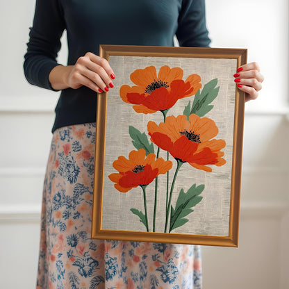 A close-up of a woman with red nail polish holding the framed artwork of three stylized orange poppies. The frame is a light brown/Orange and the print is framed with no mount or mat. The woman is wearing a dark top and a floral skirt.