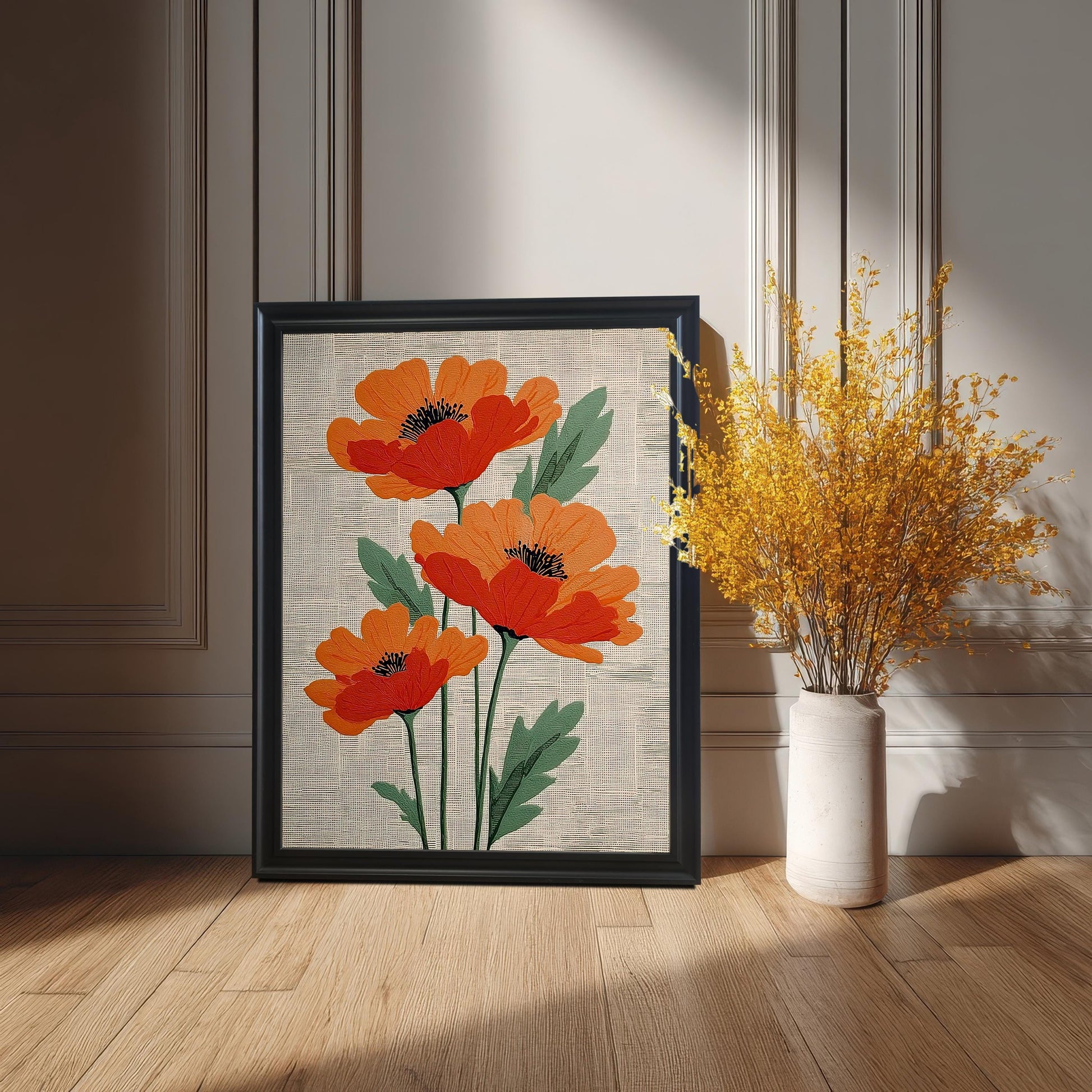 The framed artwork, featuring three stylized orange poppies, is leaning on a wood floor against a paneled wall painted a light neutral color. The frame is Black. A tall vase with a large bouquet of dried yellow flowers sits to the right.