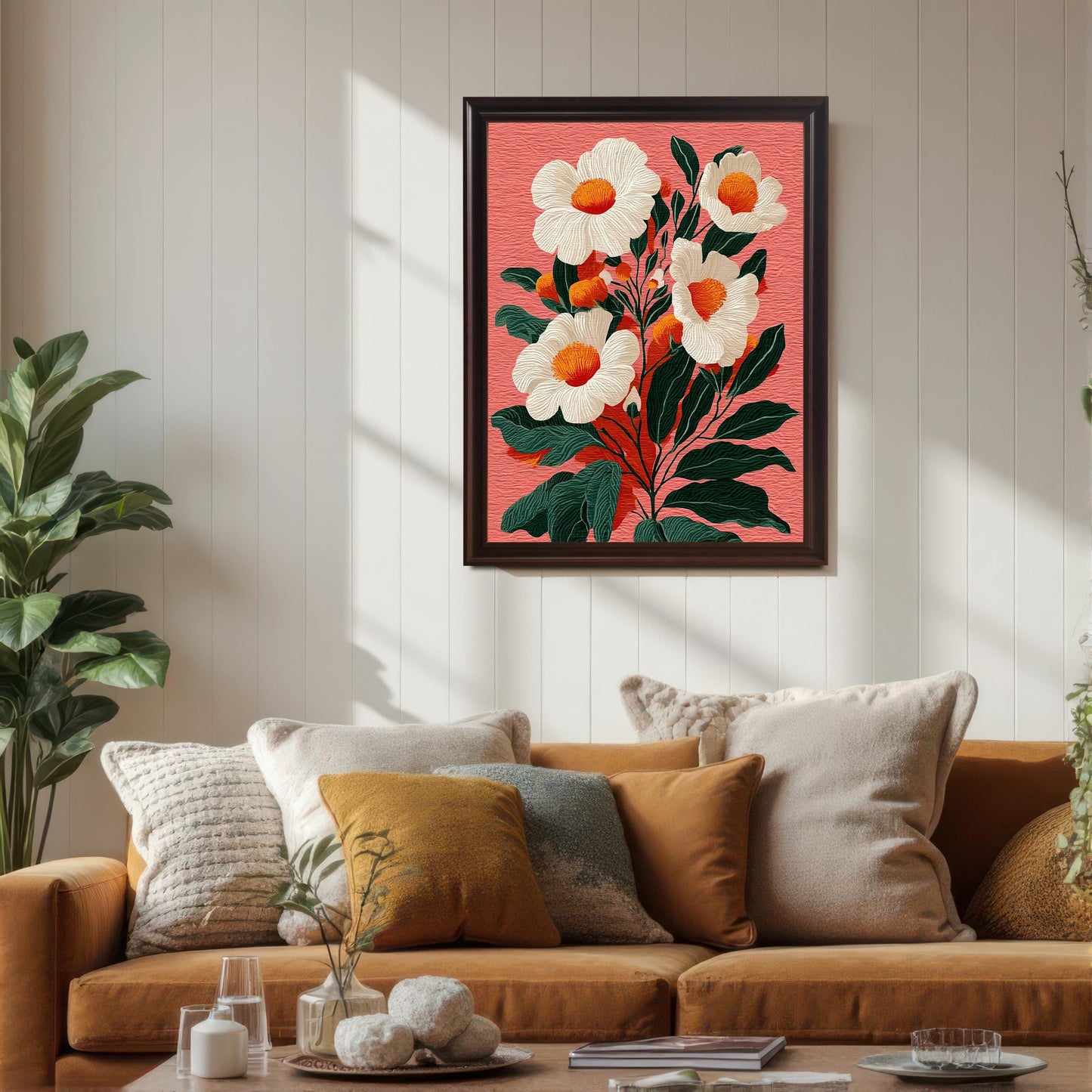 A framed floral print featuring white flowers with orange centers on a textured salmon-pink background, hanging above a mustard-colored sofa. The frame is a dark brown (Coffee) color. Sunlight casts strong shadows across the white paneled wall and the art, enhancing the cozy living room scene with pillows and a side plant.