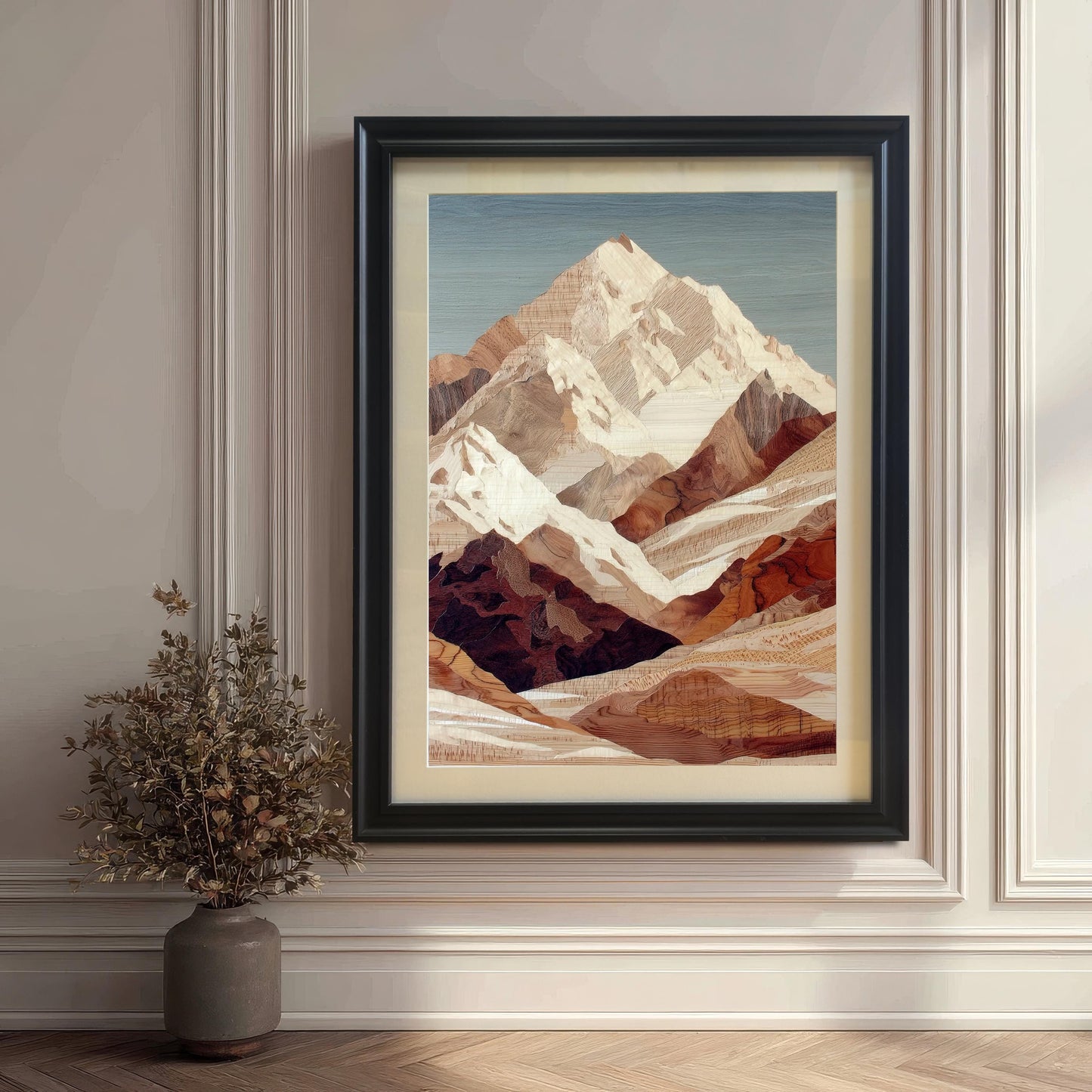 A framed mountain art print is leaning on the floor against a classic white wall with wainscoting. The frame is black and includes a light-colored mount. A small vase with dried foliage sits to the left.