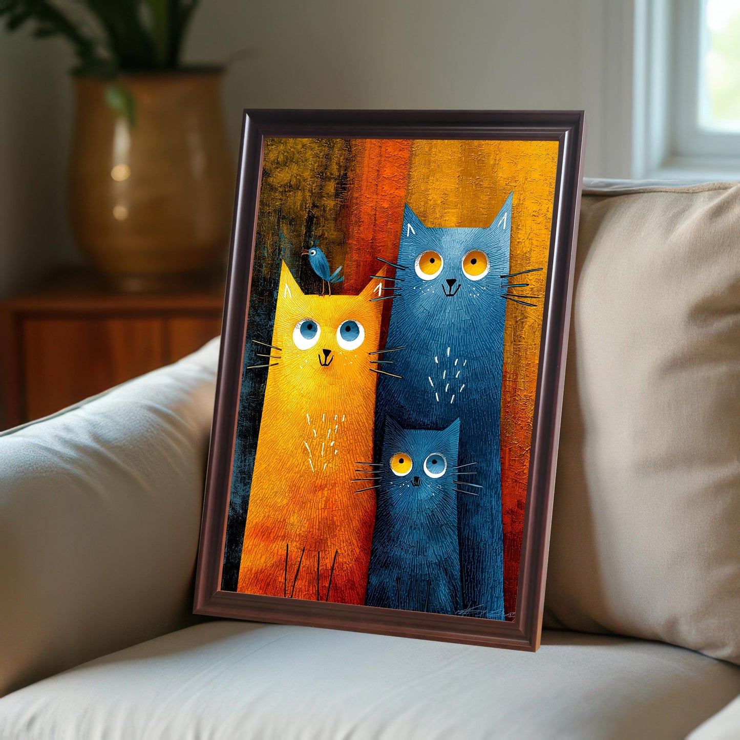 Framed print of a colorful, stylized painting of a family of three cats (one large orange cat with a small blue bird on its head, one large blue cat, and one small blue cat) leaning on a light-colored couch in a cozy, sunlit room. The frame is dark brown or coffee-colored.