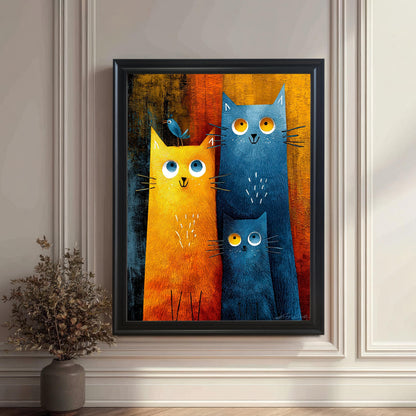 A close-up view of the cat family art print hanging on a classic white wall with wainscoting. The frame is black. The print shows a large orange cat, a large blue cat, and a small blue cat. A small dried plant in a vase sits on the floor to the left.