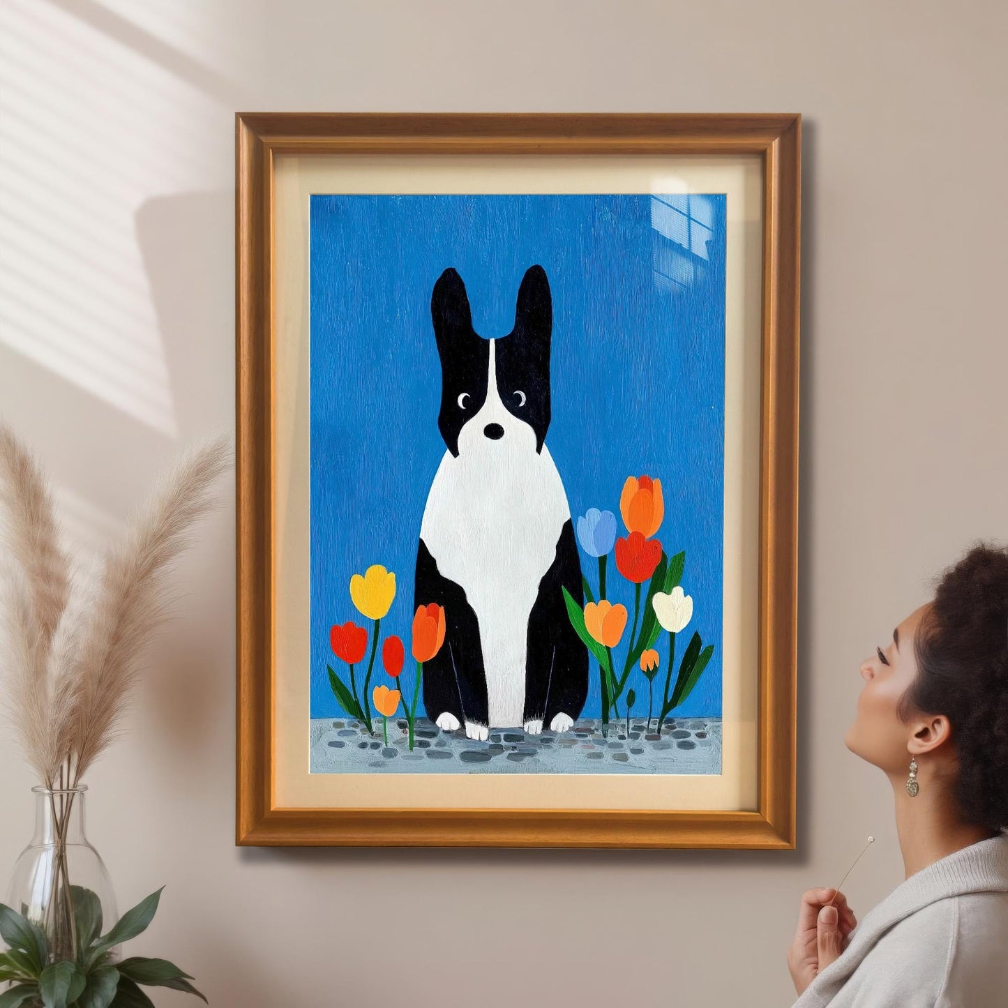 Framed art print of a black and white dog with tulips hanging on a beige wall. A woman with an afro is standing nearby, looking up at the artwork. The frame is a gold/orange color.