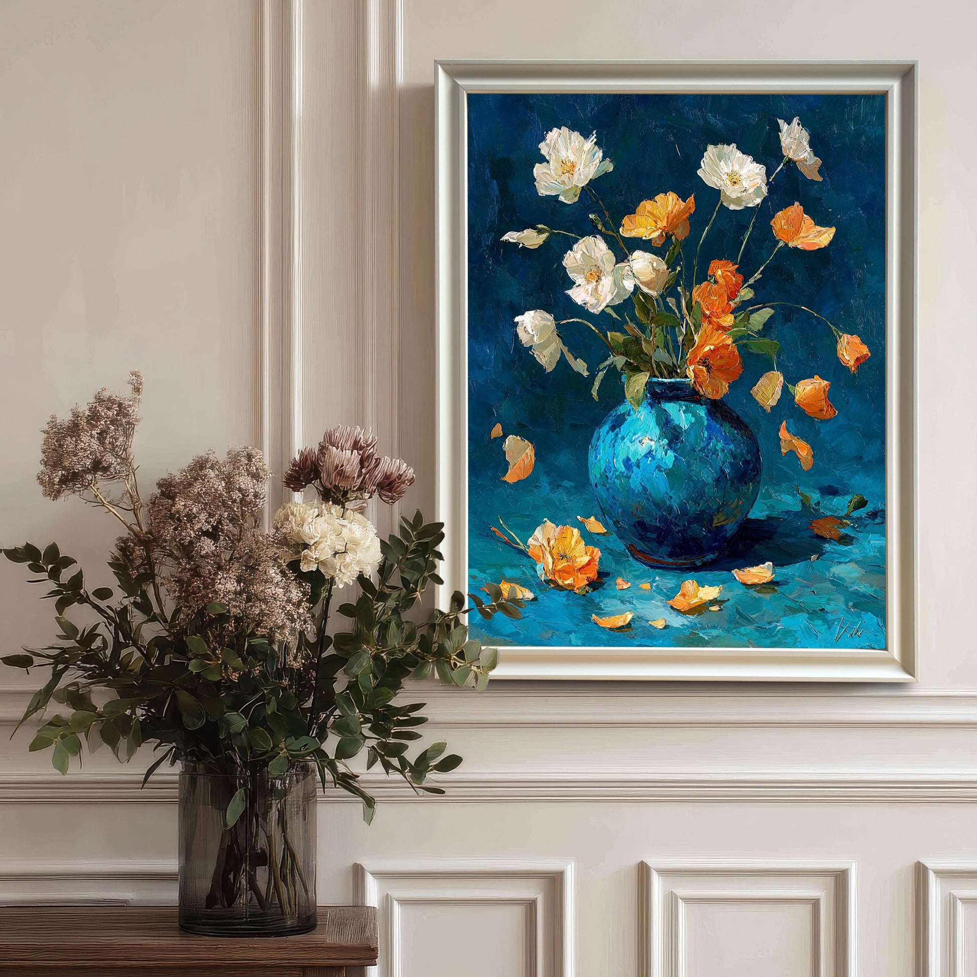 The framed floral oil painting in a White Frame hanging on a white wainscoted wall, next to a large vase of dried flowers and greenery.