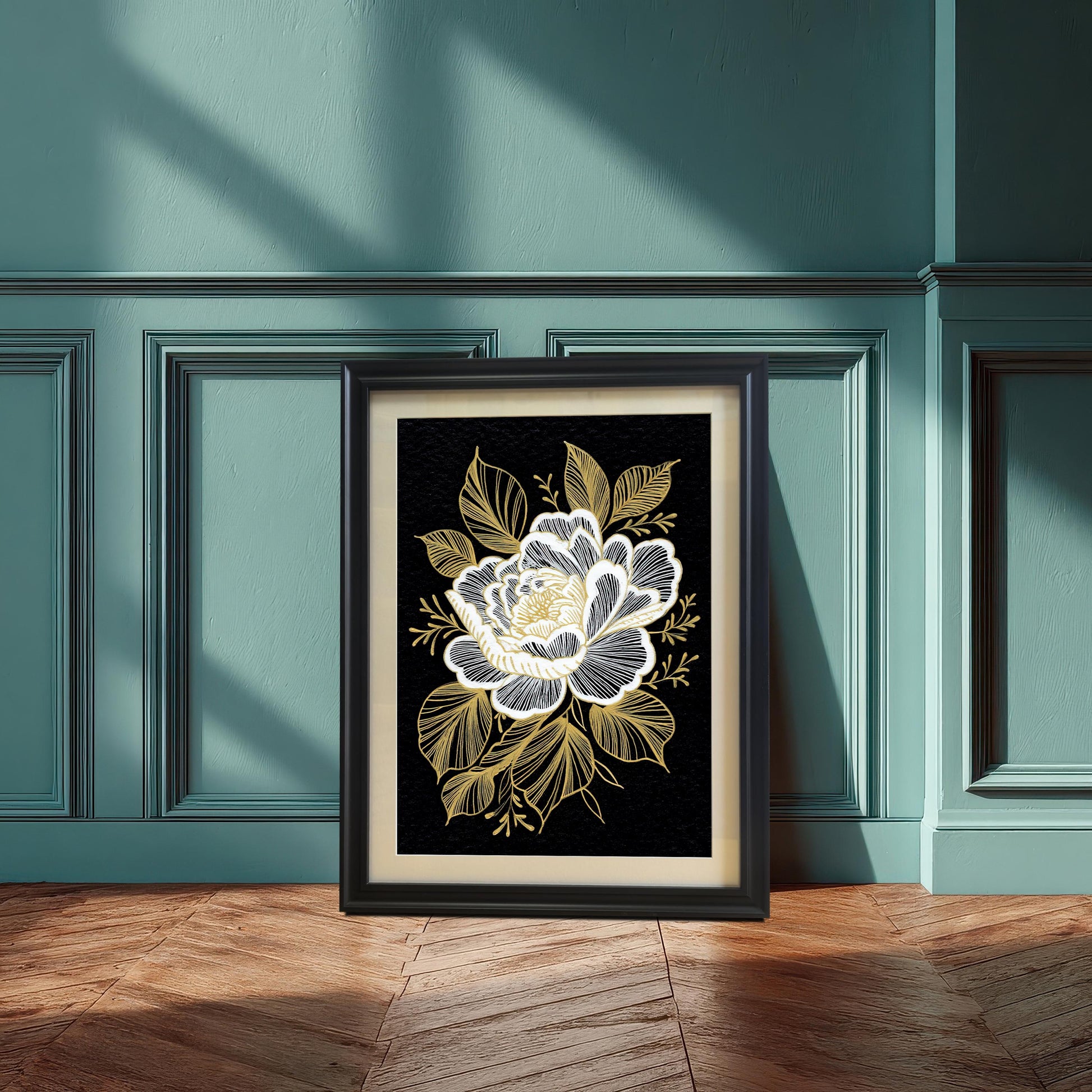 A framed black and gold floral print, featuring a white and gold stylized flower on a black background, leaning against a teal wainscoted wall and wooden floor, illuminated by a sunbeam.