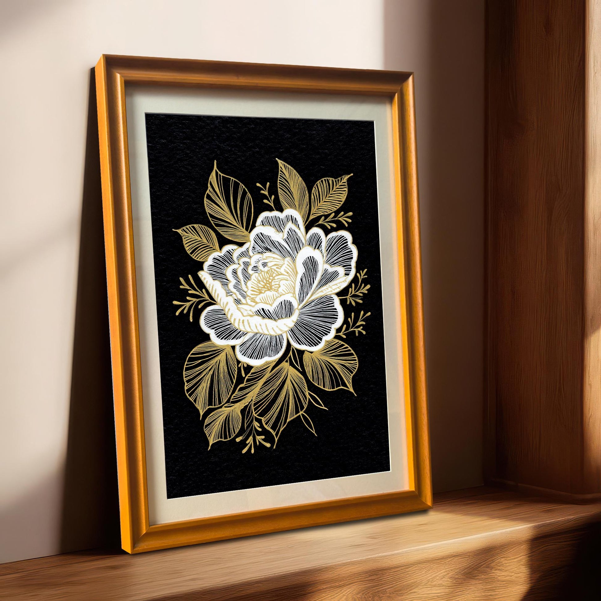 A framed black and gold floral print with a gold-colored frame, leaning on a light-colored wooden windowsill with sunlight streaming in from the right.