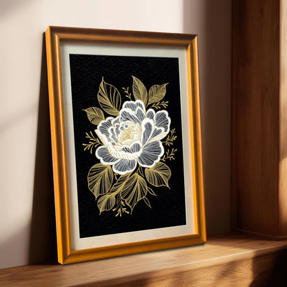 A framed black and gold floral print with a gold-colored frame, leaning on a light-colored wooden windowsill with sunlight streaming in from the right.