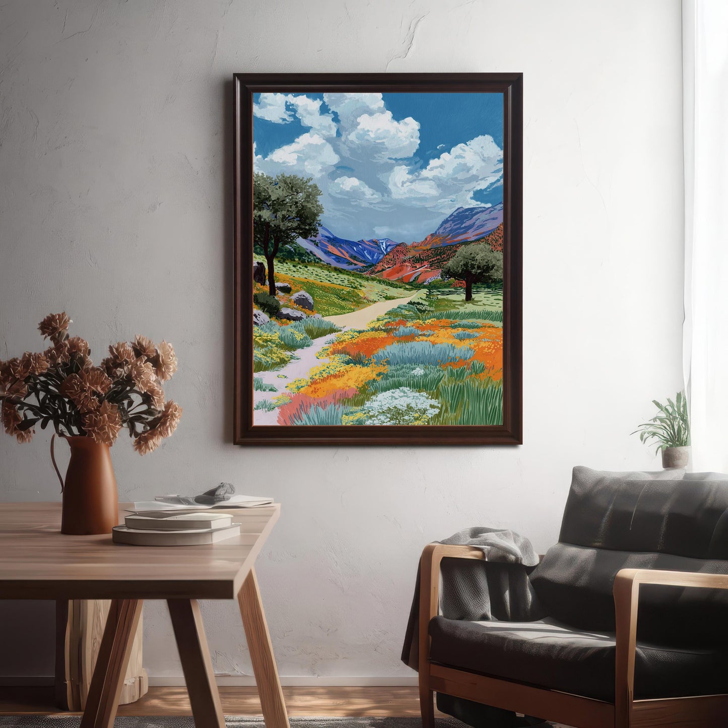 The colorful wildflower valley painting framed in a Coffee (dark brown) frame, hanging on a textured white wall above a wooden table and a dark leather armchair.