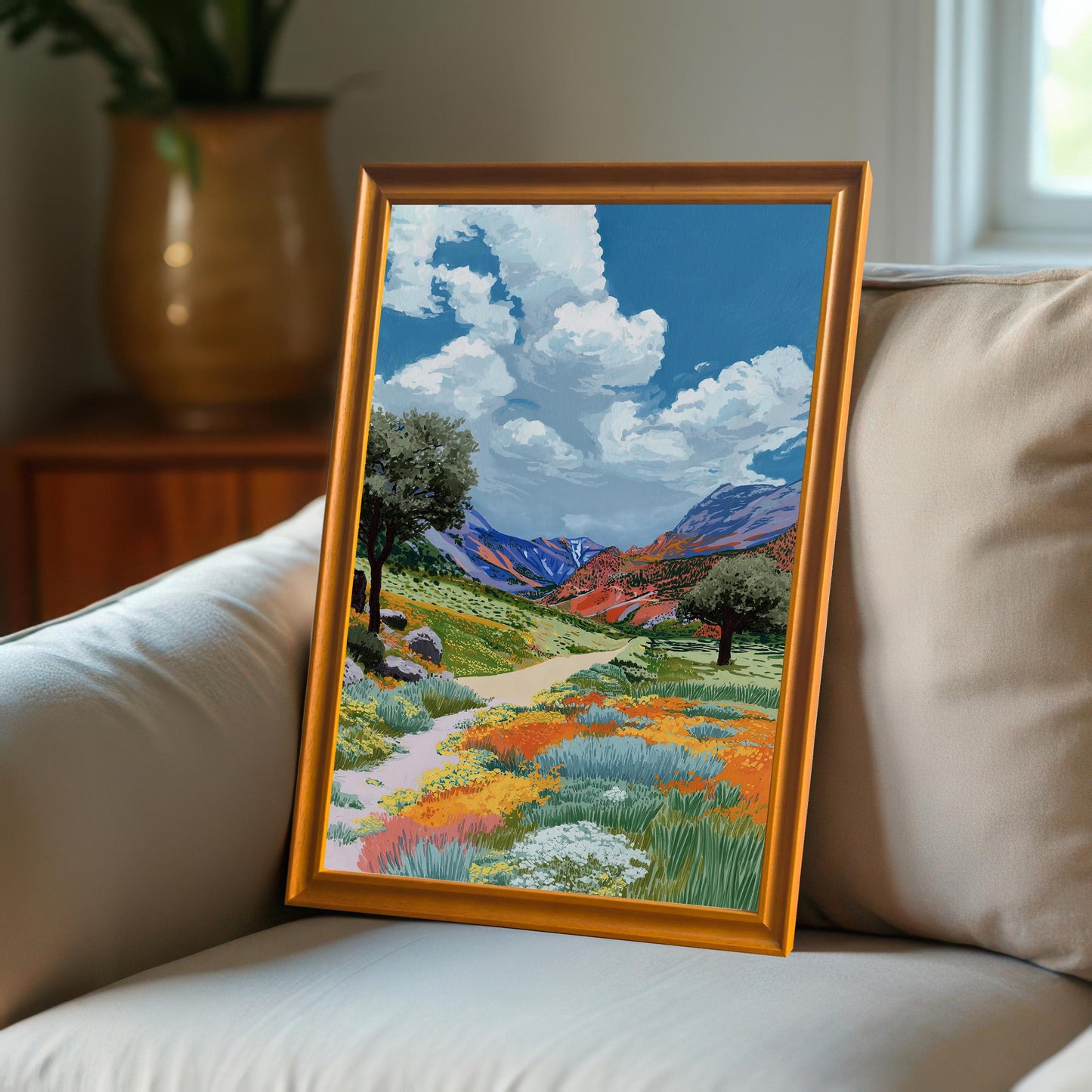 The colorful wildflower valley painting, framed in an Orange wood frame, leaning on the arm of a beige couch.