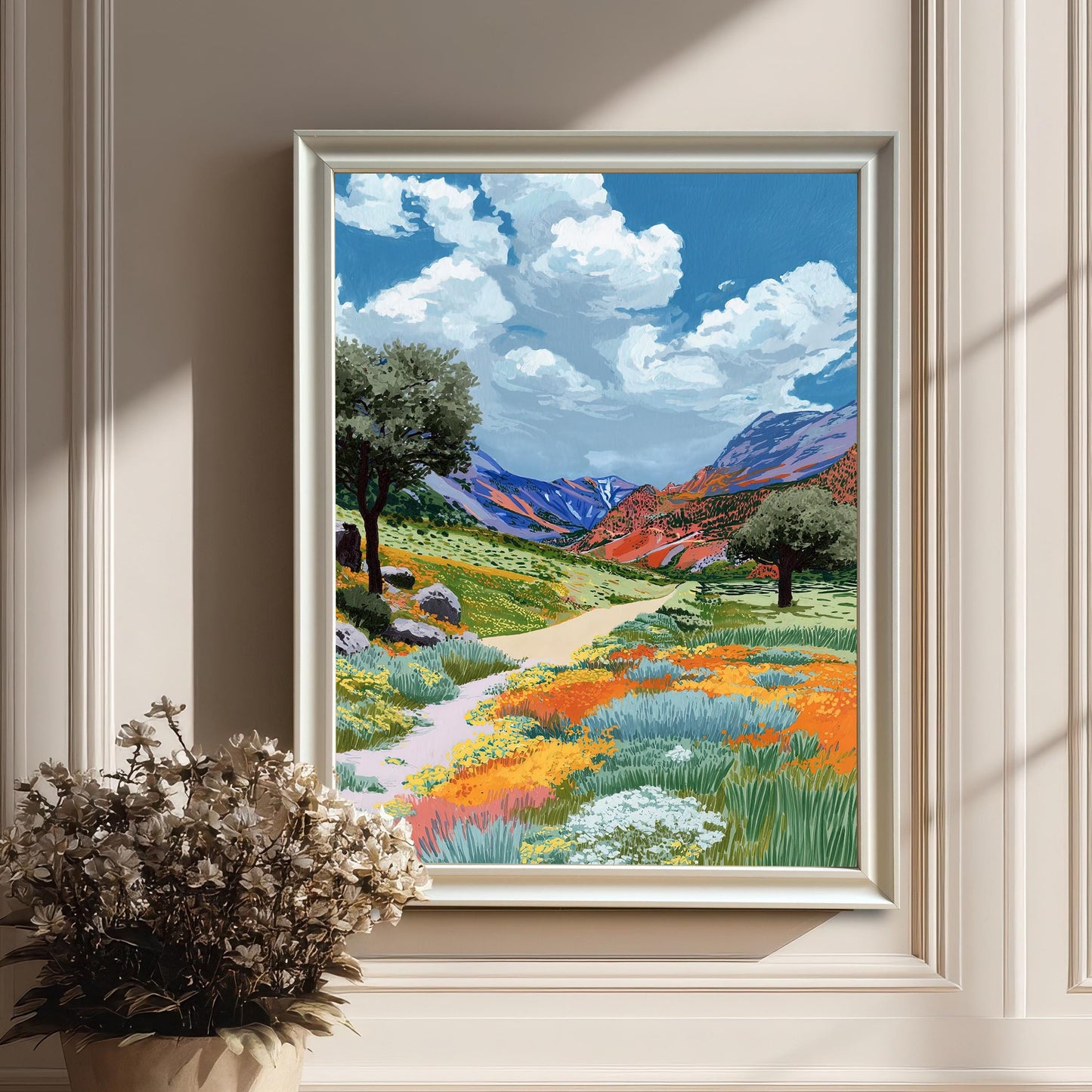 The colorful wildflower valley painting framed in a decorative white frame (no mount), leaning on a light hardwood floor against a white paneled wall. A dried floral arrangement in a vase is placed in the foreground.
