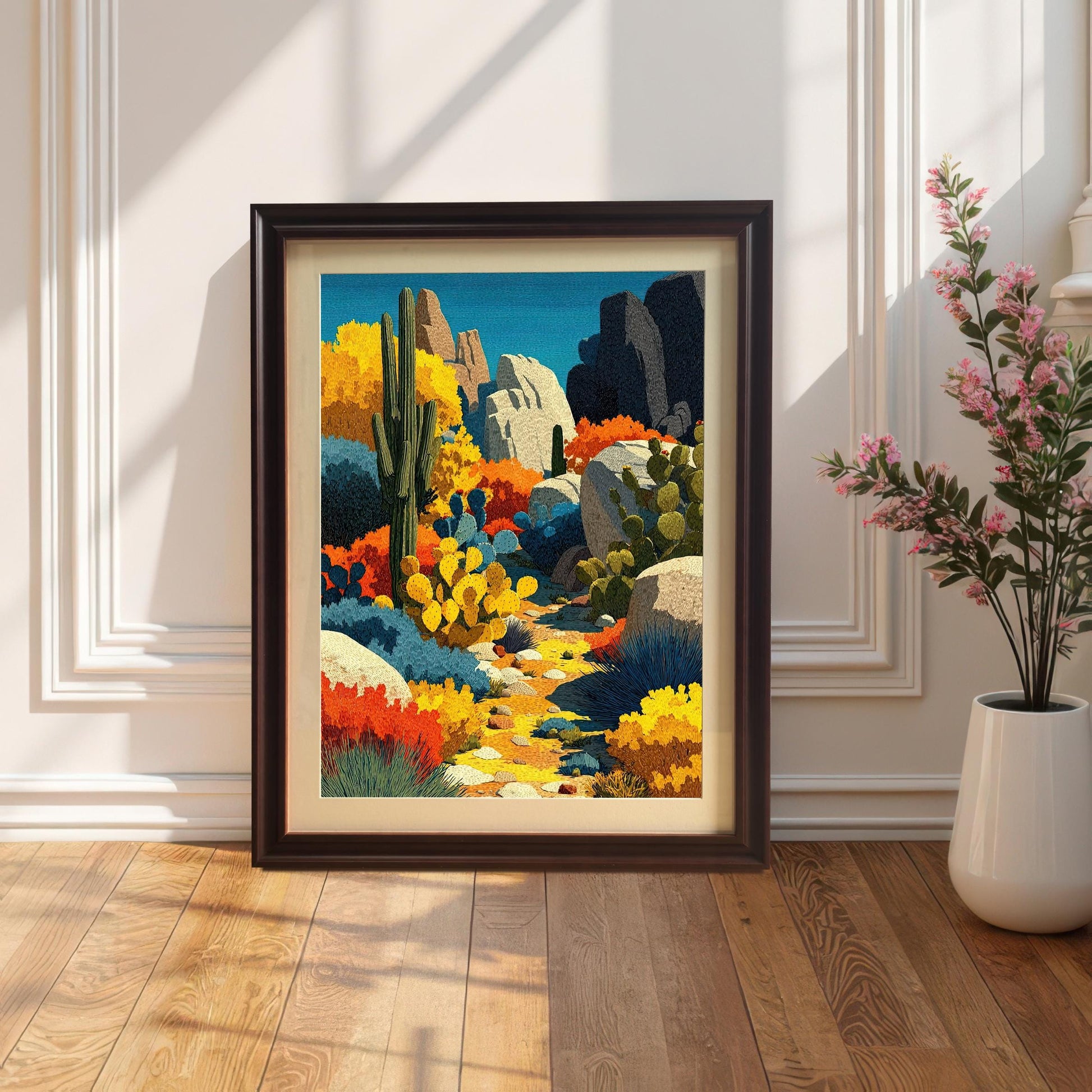 A framed, colorful desert landscape print with a dark brown (Coffee) frame and a mount, leaning against a white wainscoting wall, next to a tall white vase with pink flowers.