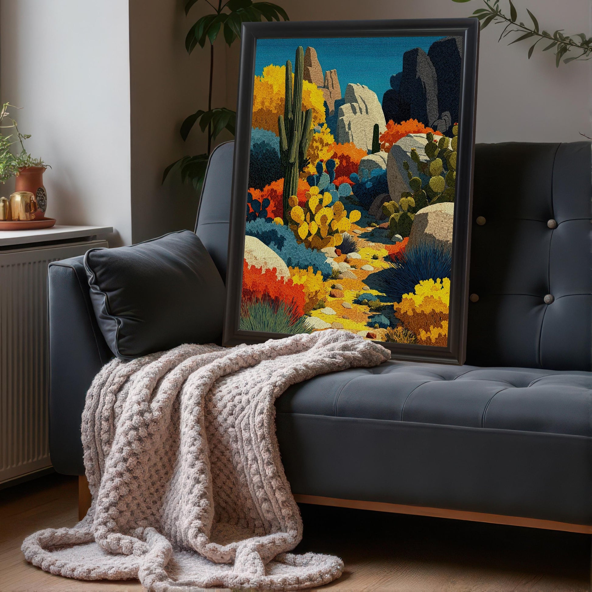 A large, black-framed, vertical art print of a colorful desert landscape leaning on the back of a dark gray tufted couch, next to a cozy, chunky knit blanket.