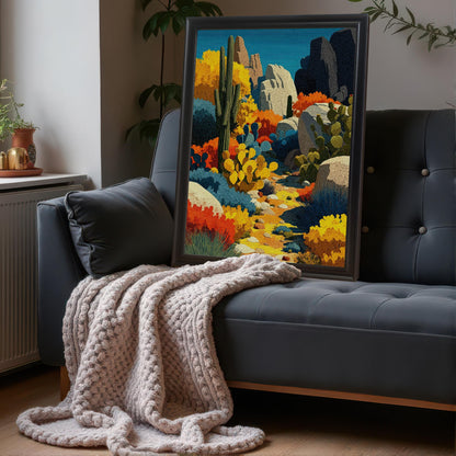 A large, black-framed, vertical art print of a colorful desert landscape leaning on the back of a dark gray tufted couch, next to a cozy, chunky knit blanket.