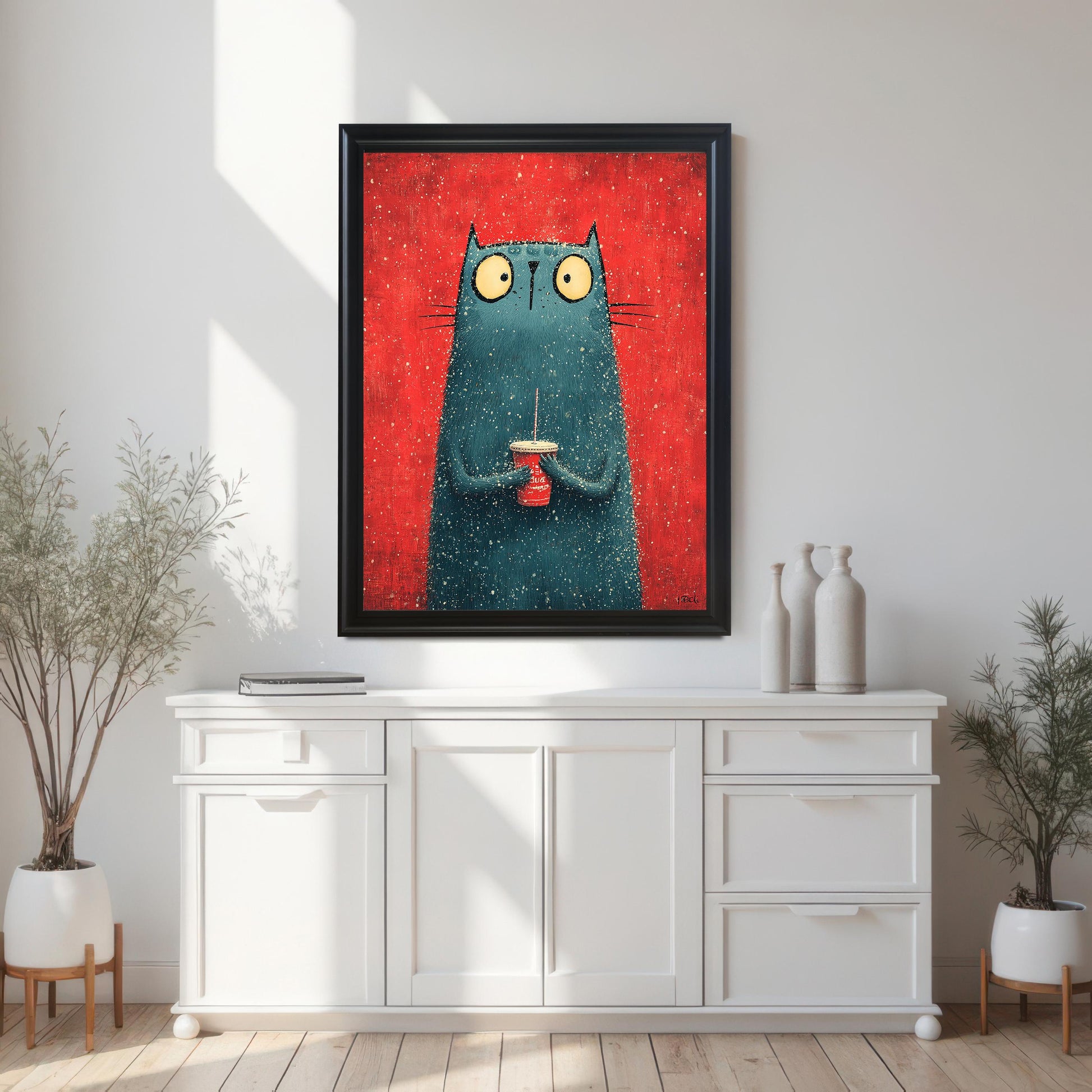 A framed art print featuring a stylized, fuzzy, blue-green cat with wide yellow eyes holding a small red cup with a straw against a bright red, textured background. The print is in a Black Frame and is centered above a long, white console table.
