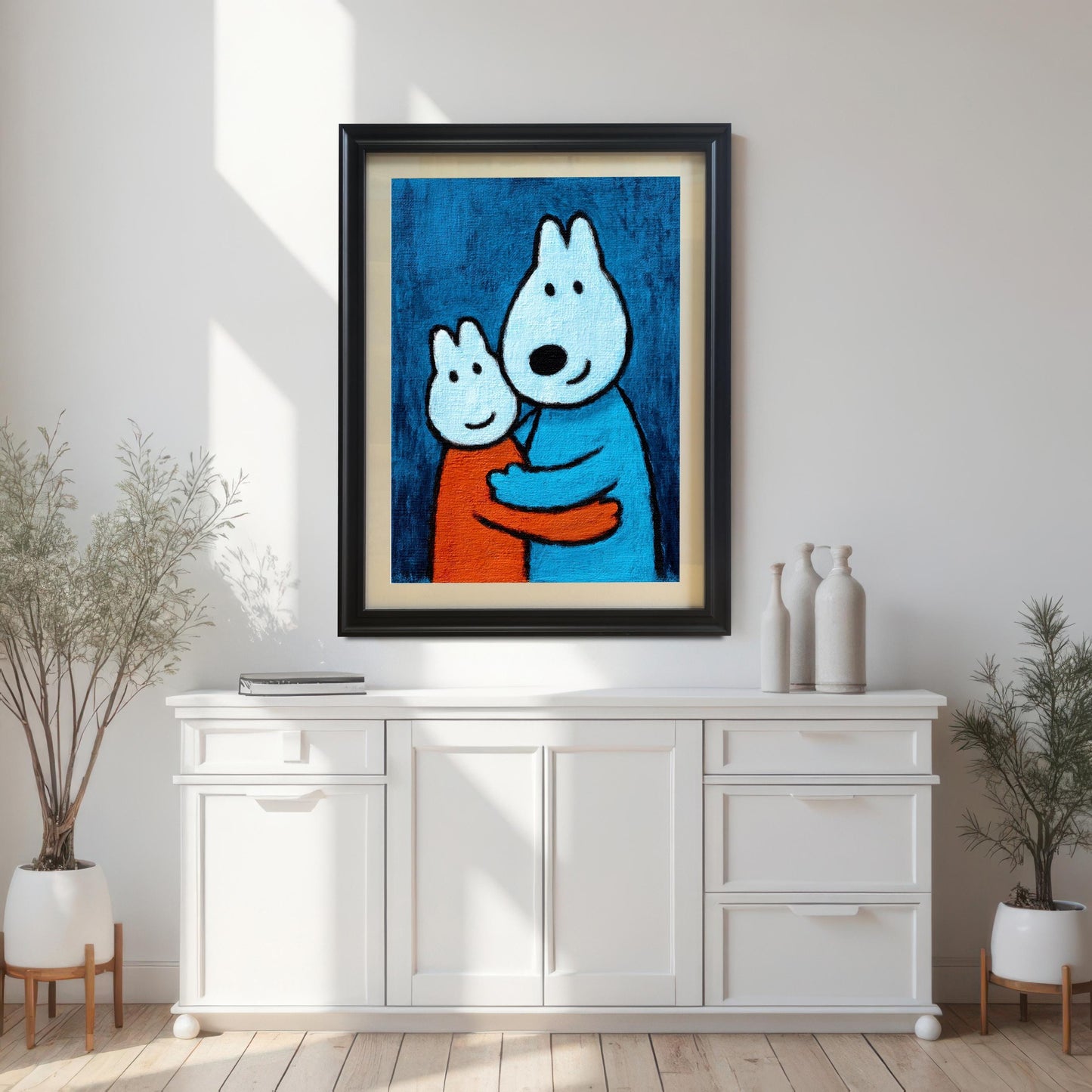 A framed art print in a Black wood frame with a mount hanging above a white sideboard cabinet in a brightly lit room with houseplants. The print features two hugging cartoon animals.