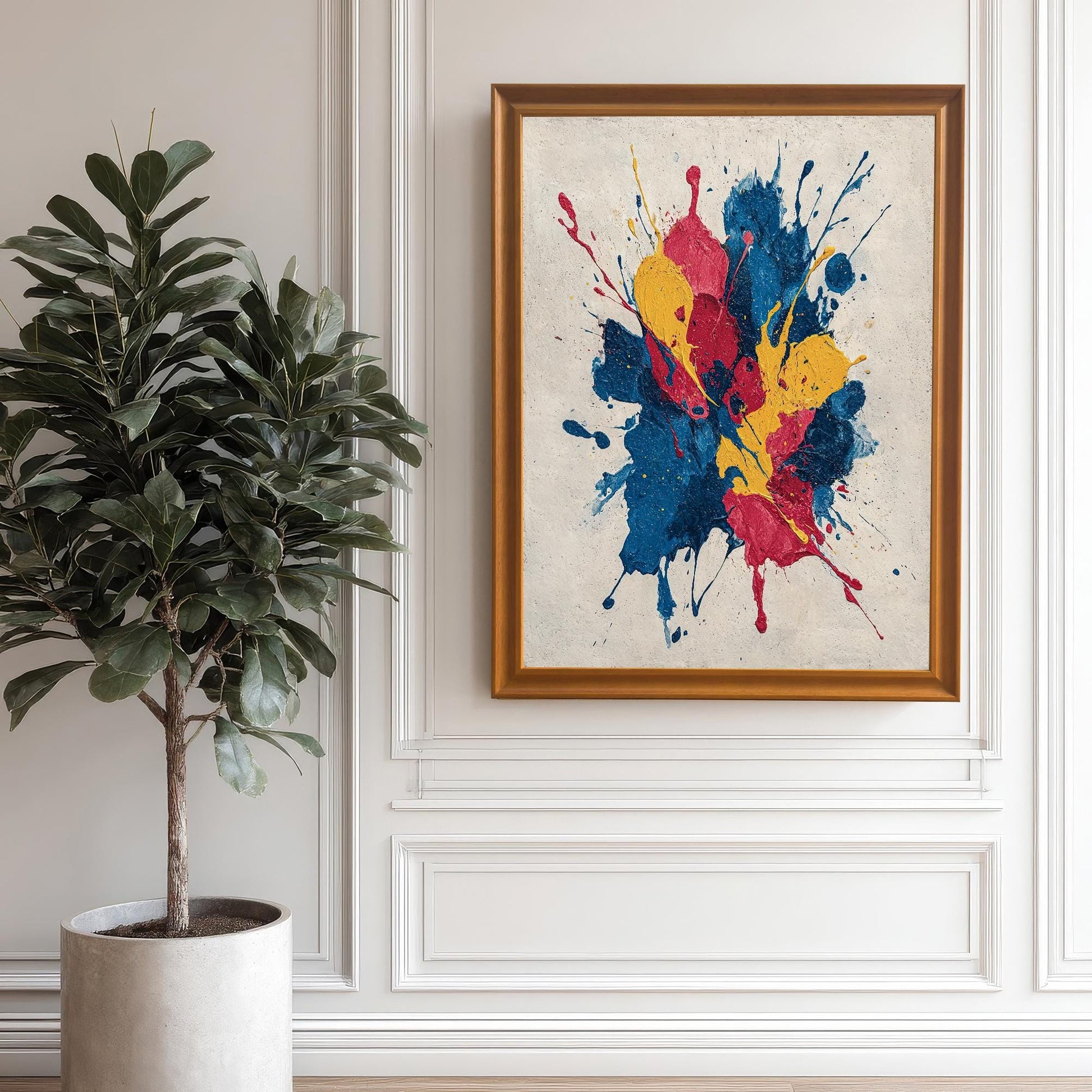 Abstract paint splatter art print (blue, red, yellow) framed in Orange/Gold/Wood Tone and hanging on a white wall with classic wainscoting paneling. A large indoor plant is positioned to the left.
