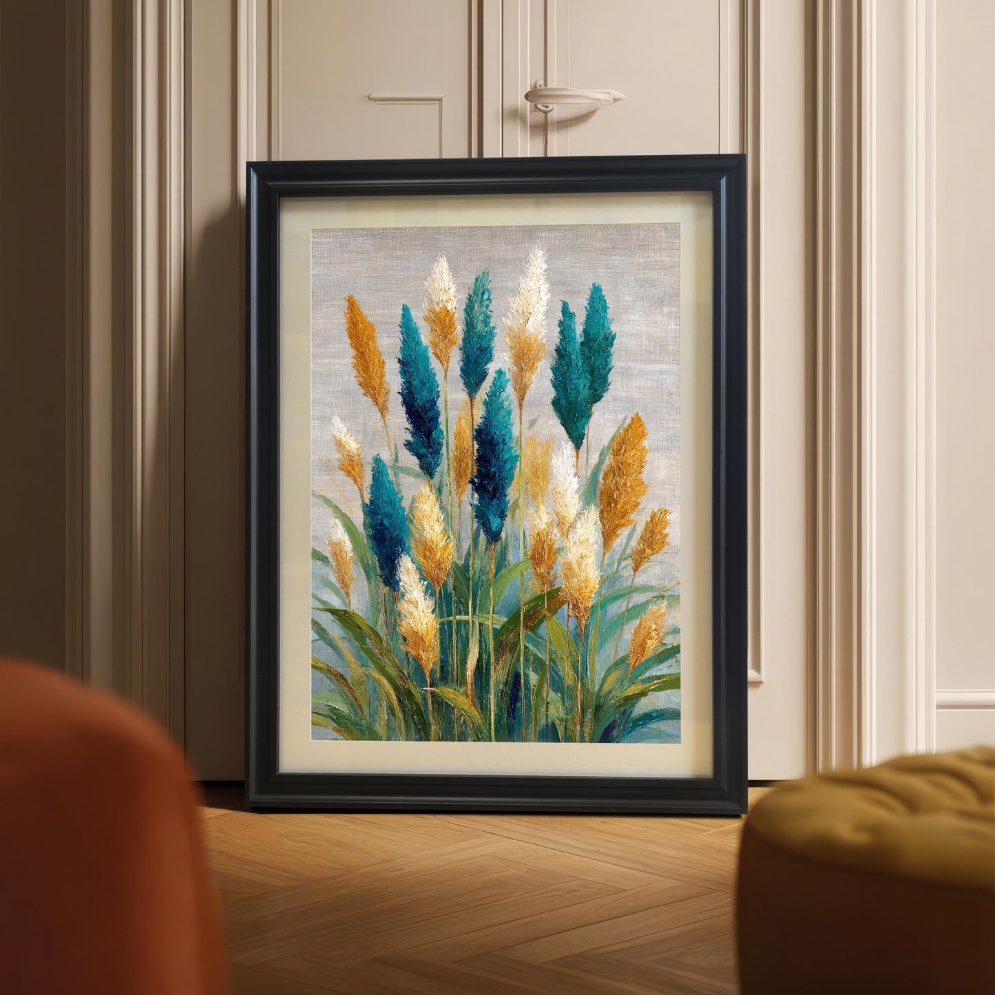 Black-framed art print, propped on a light wood floor against a beige, paneled wall or cabinet. The print features a modern oil painting of colorful pampas grass plumes in teal, gold, and white. A mustard yellow and orange upholstered piece of furniture is visible in the foreground.