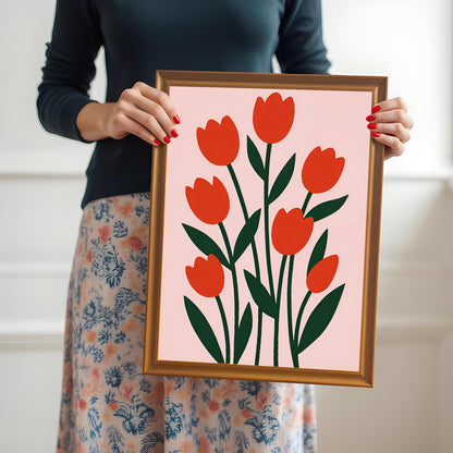 A person with red painted nails and wearing a floral skirt holds up a gold-framed print of red tulips on a light pink background at chest level.
