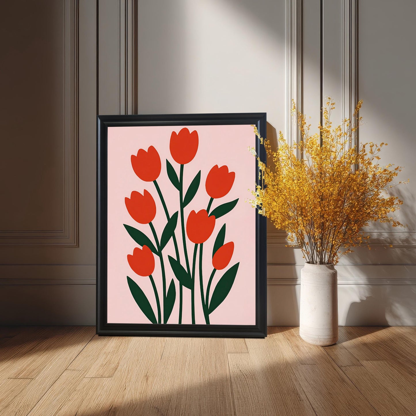 A black-framed print of the red tulip art rests on a wooden floor next to a white vase holding a bouquet of dried yellow flowers.