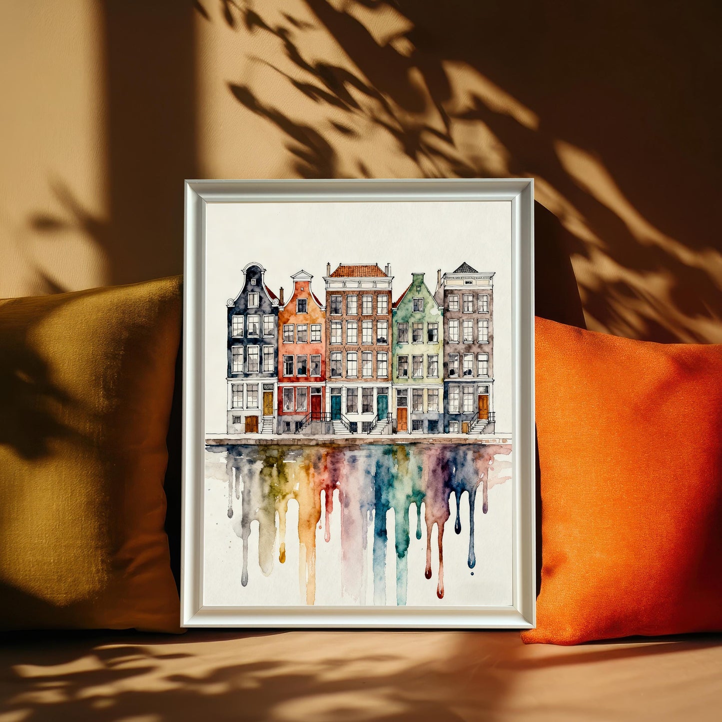 Watercolor print of colorful canal houses with a dripping rainbow reflection, framed in a white frame propped against a couch between a yellow and an orange pillow, with sunlight casting plant shadows.