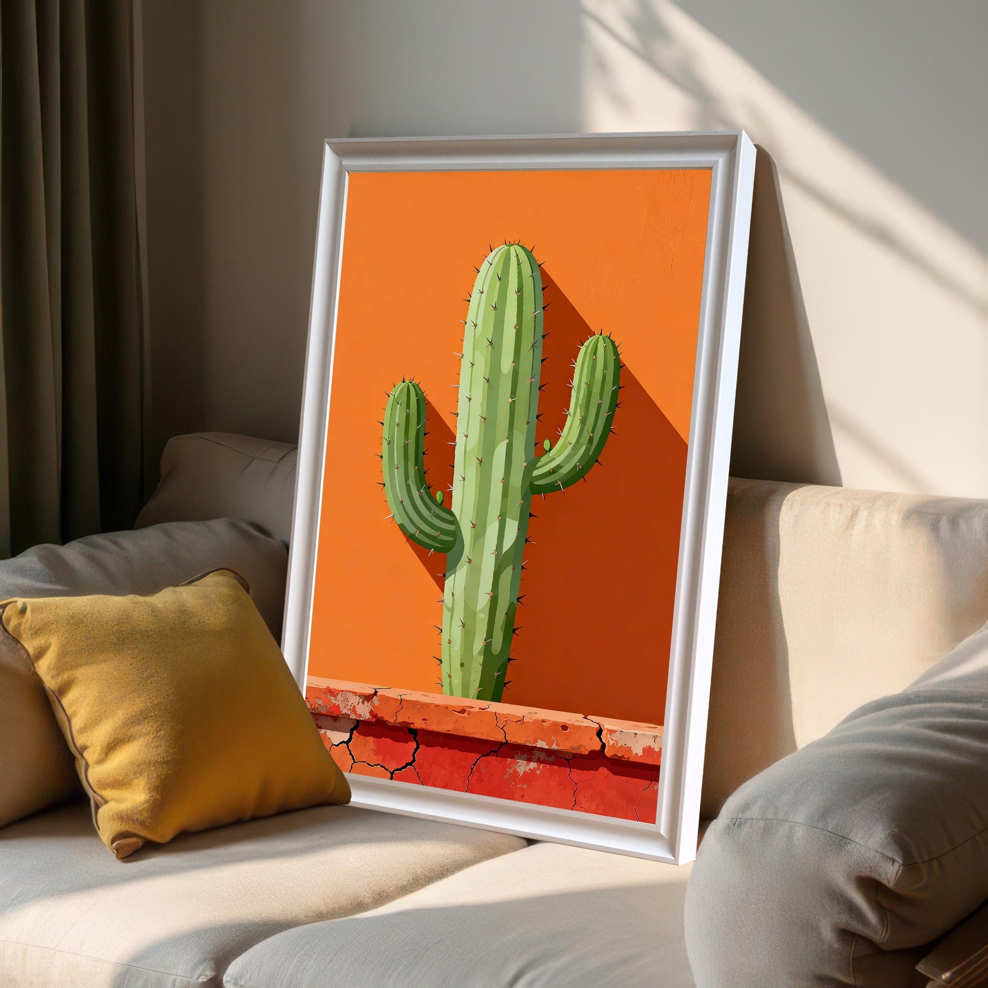 Framed cactus print in a White frame leaning on a light-colored sofa next to a yellow pillow, showing bright natural light and shadows.
