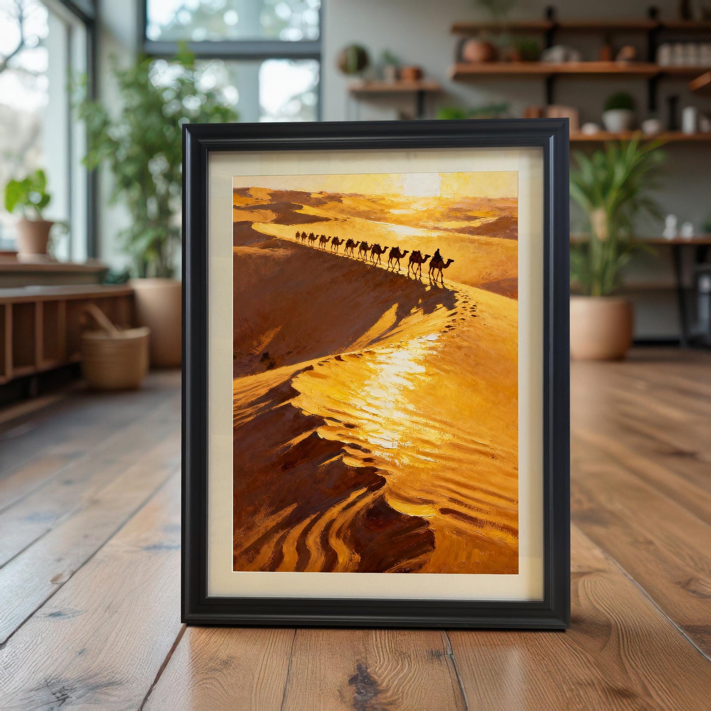 The desert camel caravan art print in a Black Frame with a Mount (mat), standing upright on a polished wooden floor. The background is a bright, modern room filled with indoor plants, giving a bohemian or nature-inspired decor vibe.