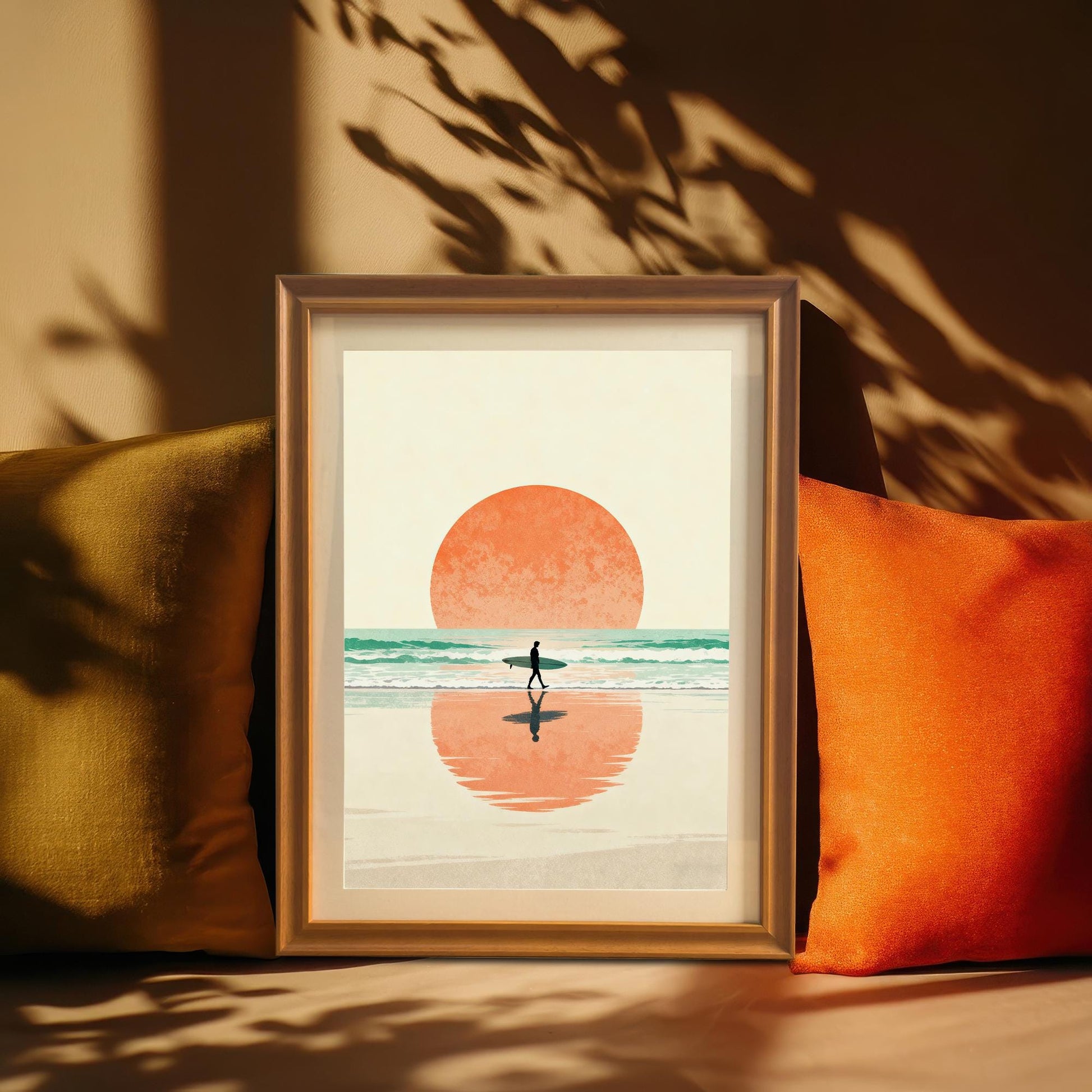 The surfer sunset art print in a warm wood frame with a Mount, standing between a gold/olive and a bright orange pillow, set against a cozy background with dramatic leaf shadows. This highlights the Boho, coastal art style.
