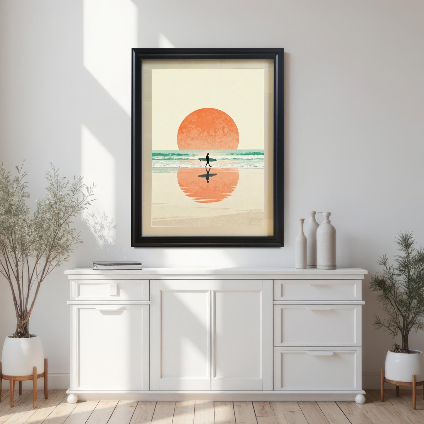 Vertical art print of a minimalist surfer walking on the beach at sunset, featuring an orange sun and turquoise water, framed in Black with No Mount. The art is hung above a white console table, flanked by two potted plants, showcasing the print in a bright, modern room.