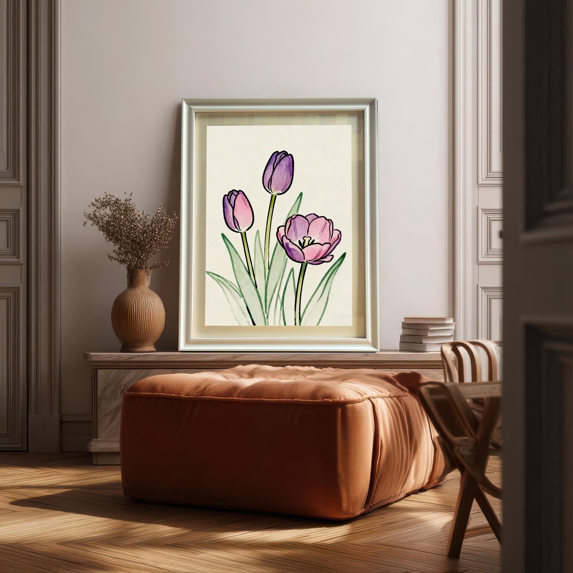 Lifestyle photo showing a framed art print of pink and purple tulips resting on a mantelpiece or cabinet, leaning against a decorative wall, positioned above a large brown leather ottoman. The frame is a light, possibly silver or natural wood, color.