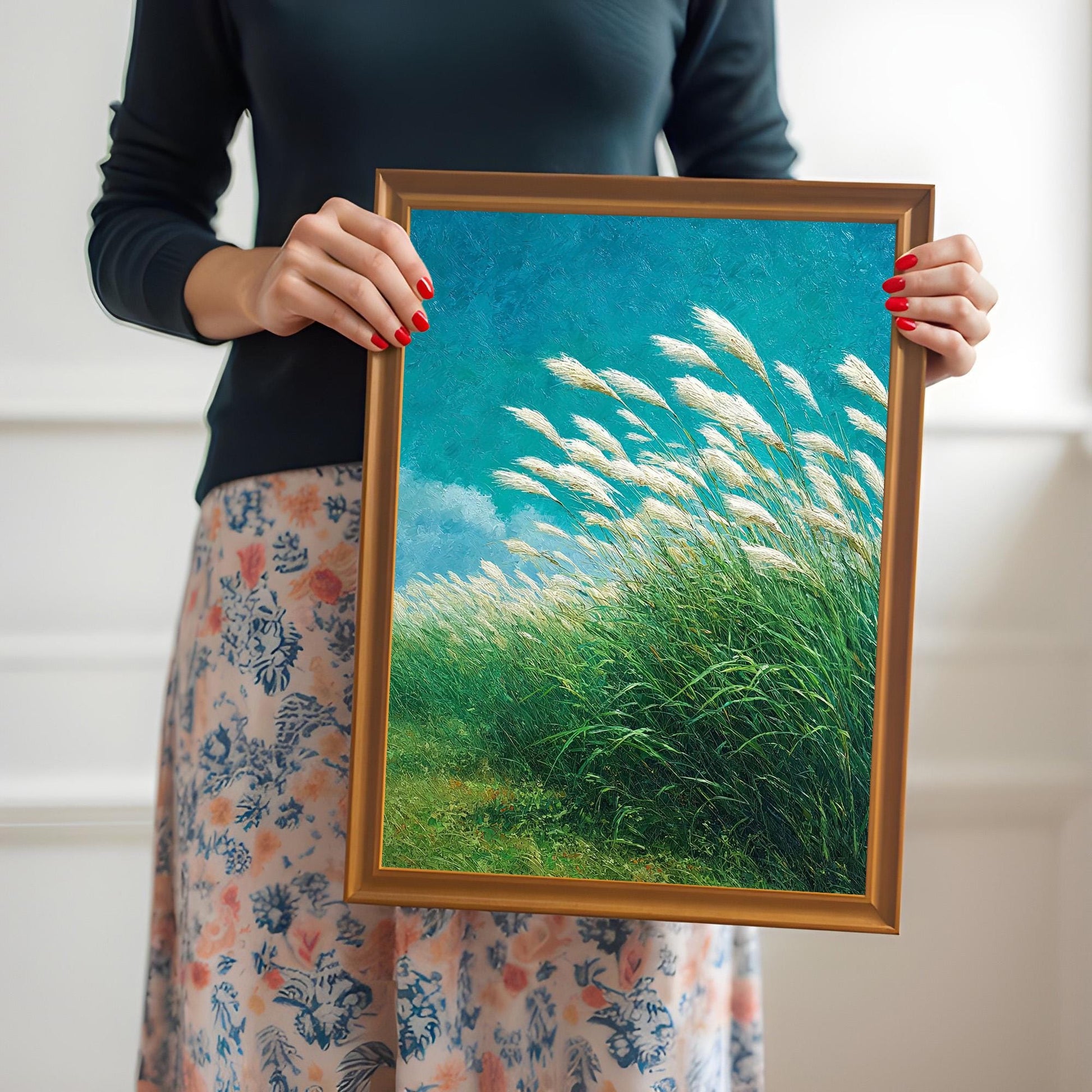 Close-up photo of a woman holding a framed art print of white pampas grass. The frame is a natural wood color (Orange option), and the woman has red painted nails and is wearing a dark top and a floral skirt.