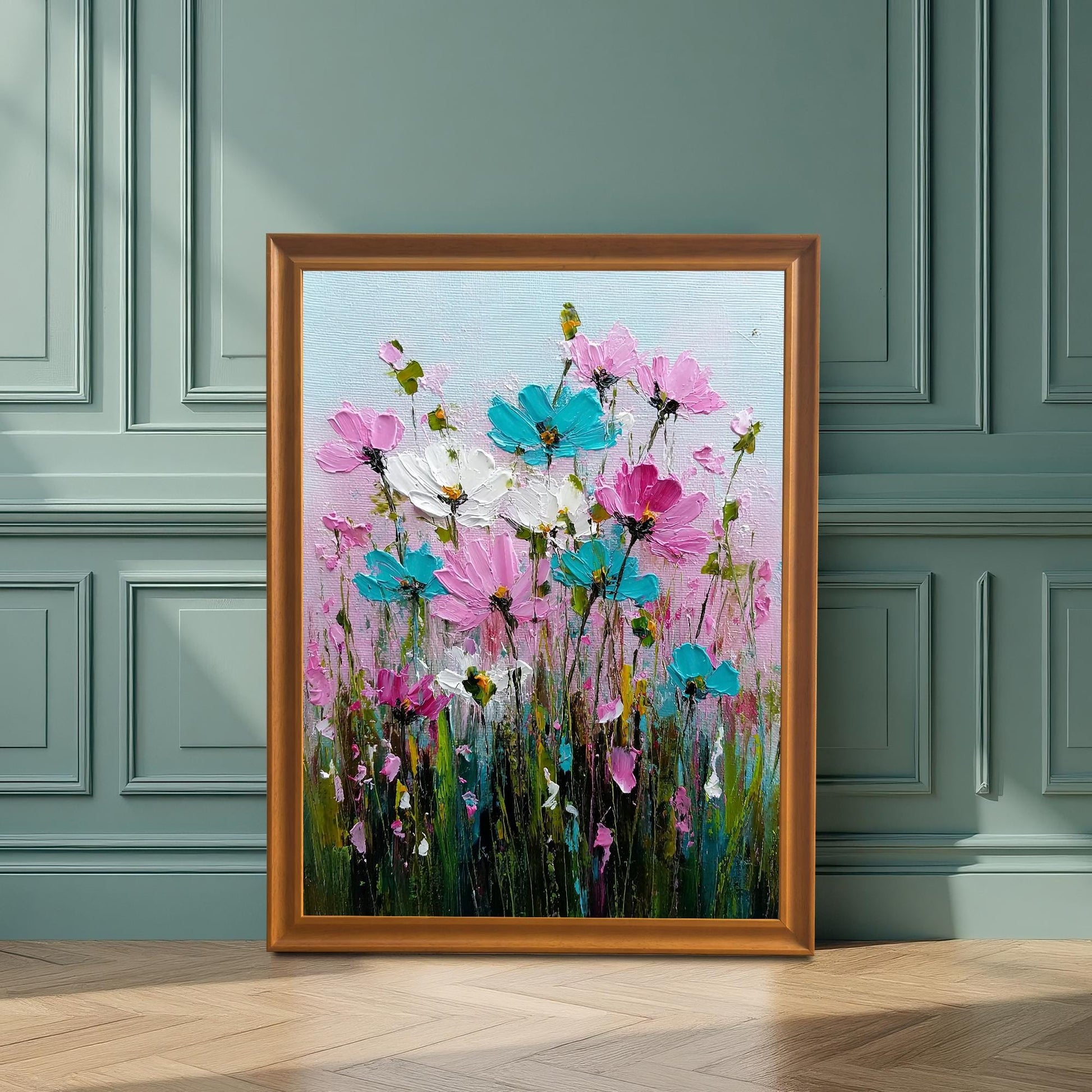 Orange-gold framed art print of a vibrant, abstract field of pink and turquoise flowers. The framed art is leaning on a wood floor against a decorative, sage-green paneled wainscoting wall with a strong shadow cast on it.