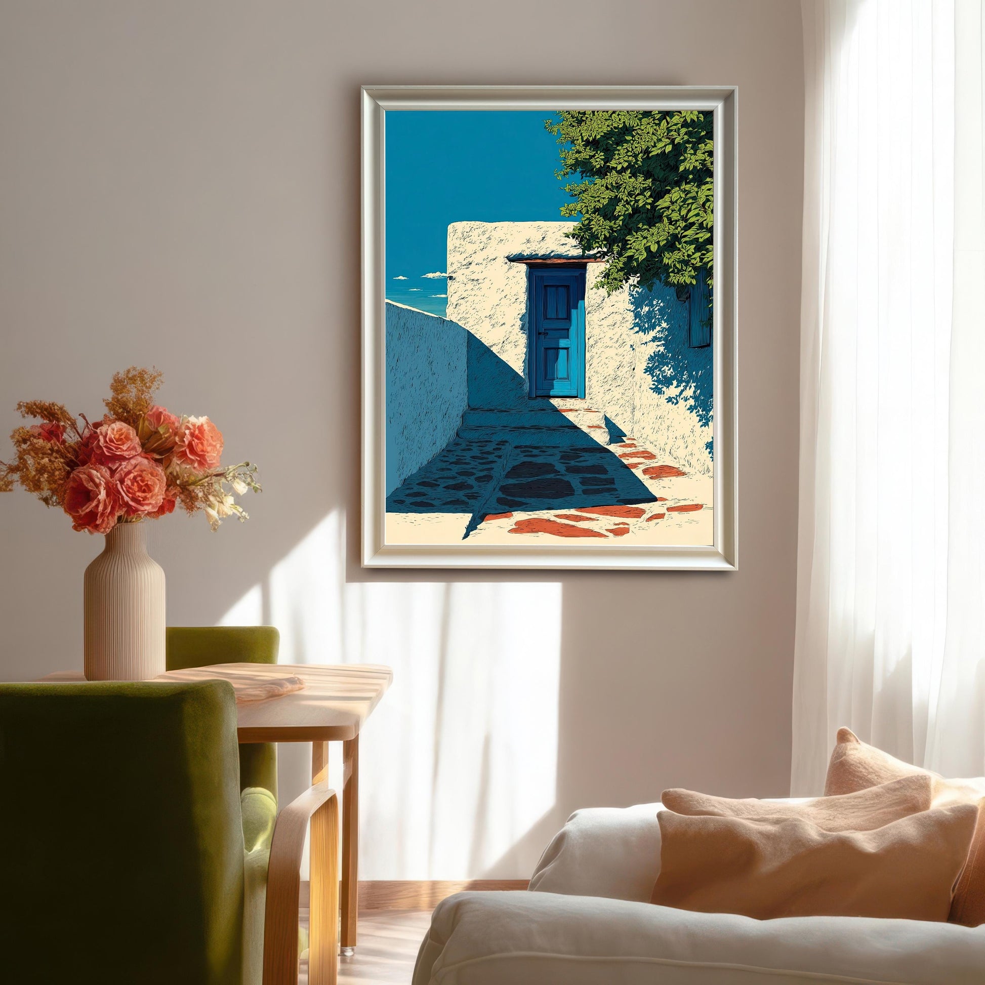 A large, framed art print of a bright white building with a blue door and green foliage, hanging in a sunny dining room above green chairs and a white couch. The frame is silver/light gray.