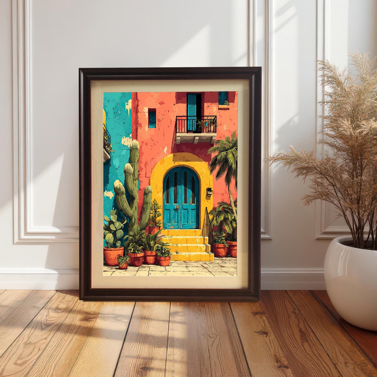 Bright, colorful print of a Mexican-style house with a yellow arch and blue door, framed in a dark Coffee (brown) frame with a mount/mat. The framed art is resting on a wooden floor against a white wainscoted wall next to a large potted plant.