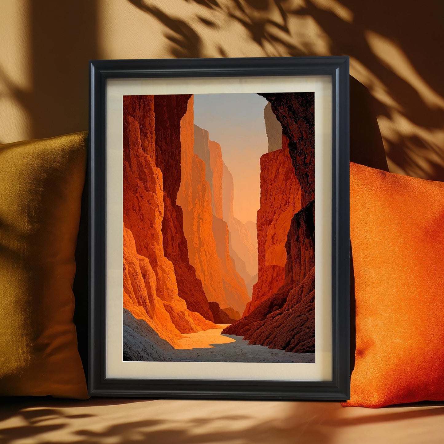 Framed canyon art print (Black frame with a mount) resting on a couch between two pillows (mustard yellow and bright orange). The image shows tall, textured red-orange canyon walls fading into the distance. A dramatic shadow from a window frame falls across the background.