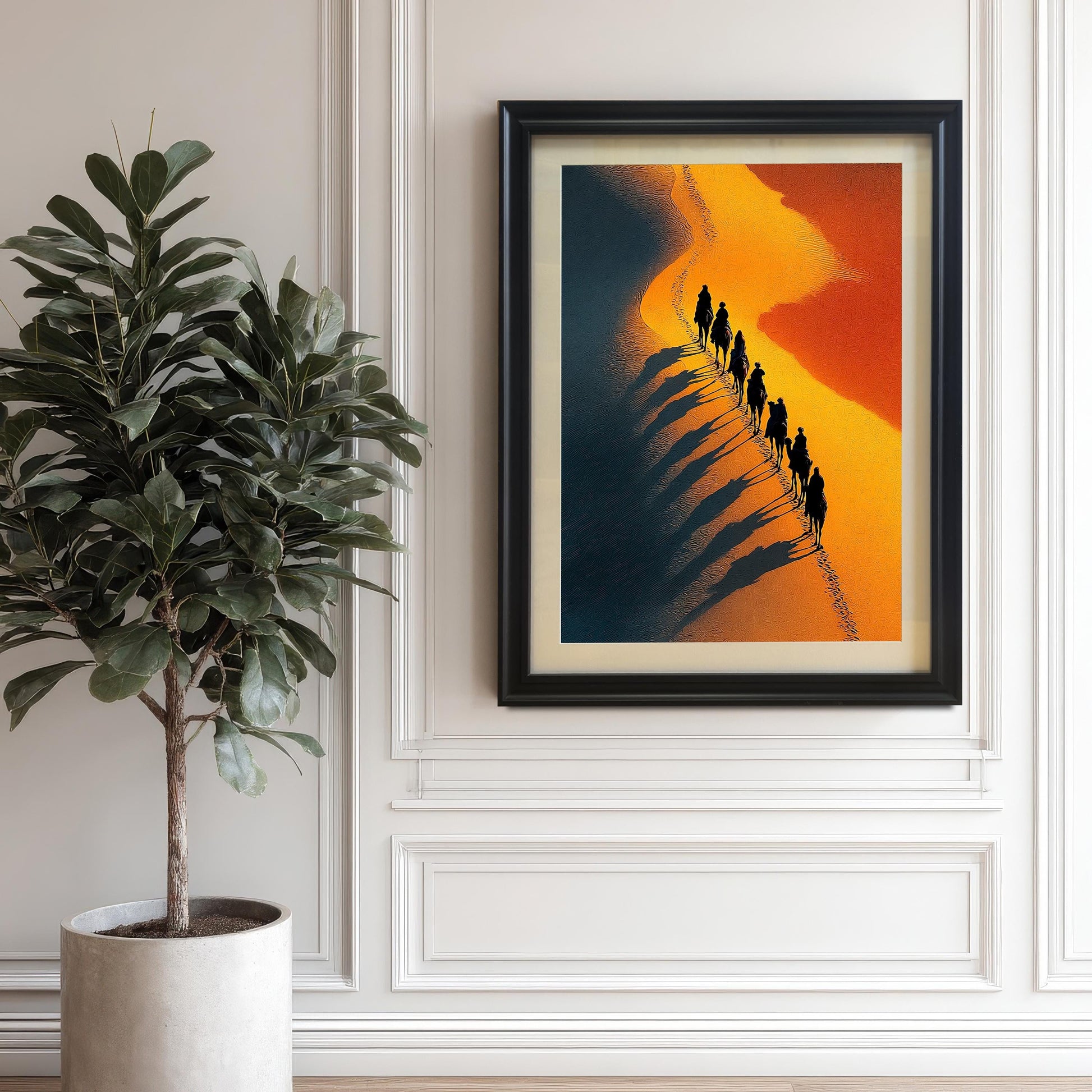 Photo of the framed art print (Black frame with a mount) hanging on a light, paneled wall next to a large potted plant. The art shows a high-contrast desert scene with a line of camels and dramatic, exaggerated shadows