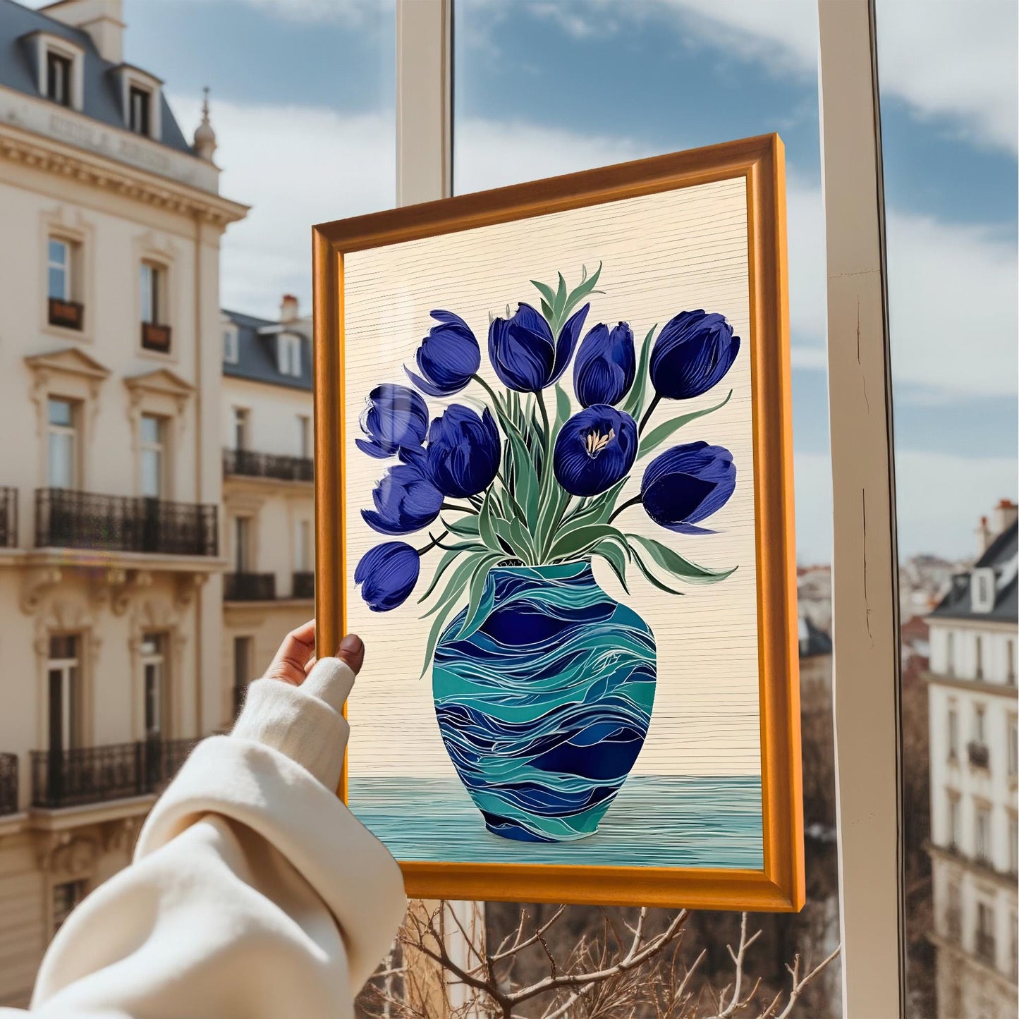 A hand in a cream sweater holds up a framed art print of blue tulips in a teal vase against an exterior window view of a European city. The frame is Orange-colored wood.